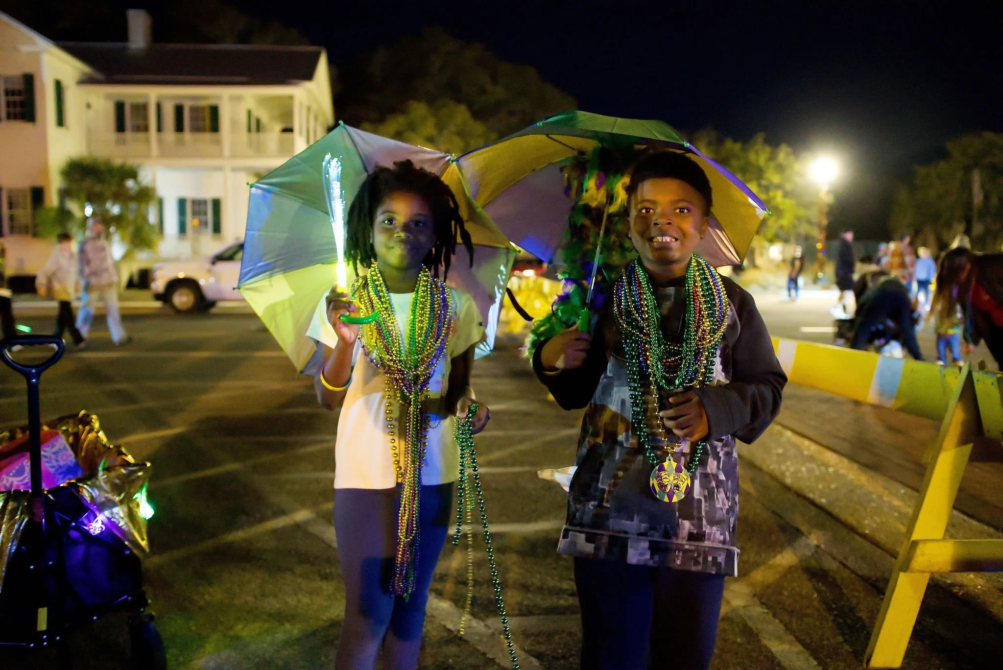 Two children smiling and wearing Mardi Gras beads, holding umbrellas, in a street at night during a celebration, with houses and other people in the background.