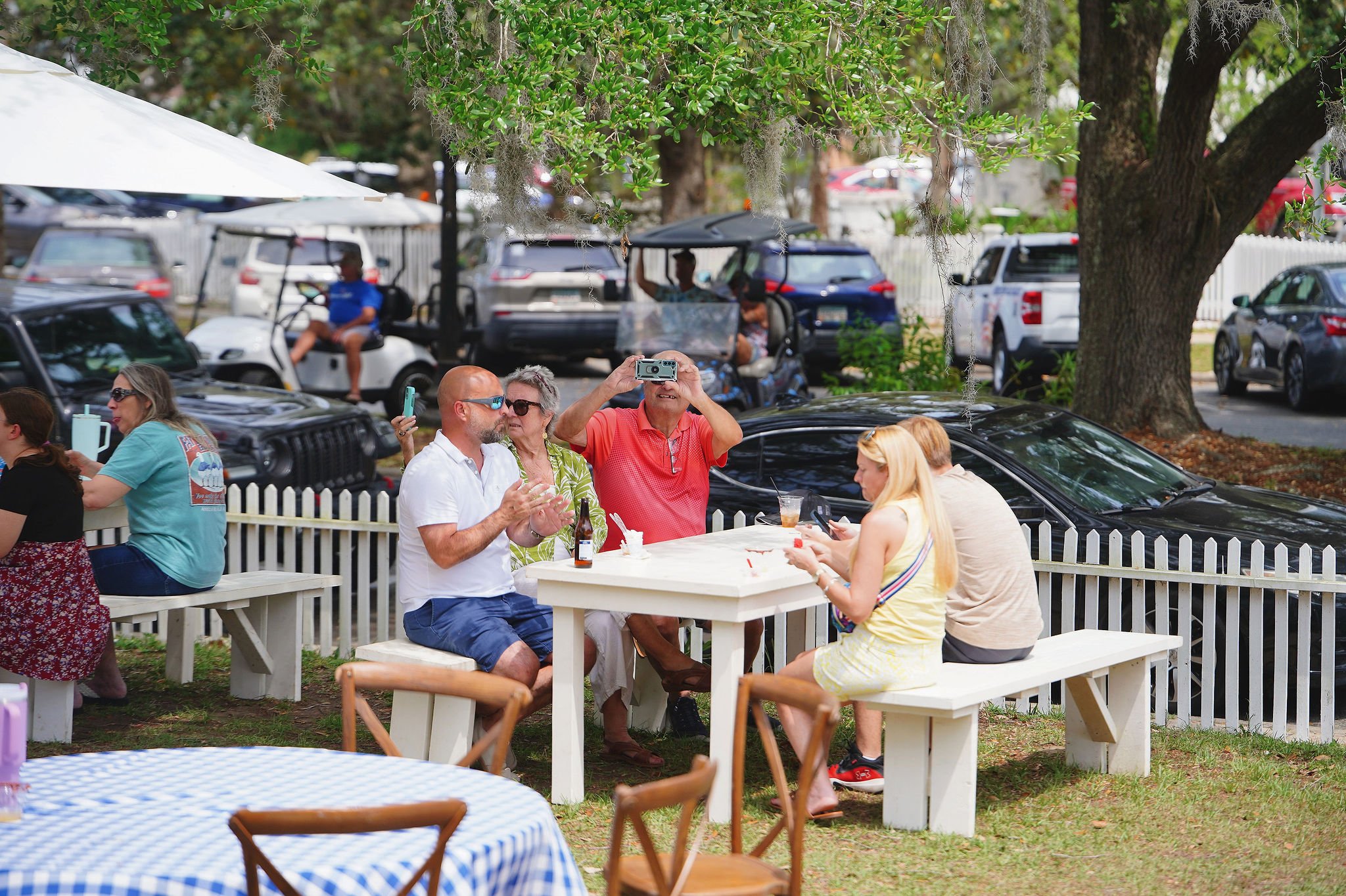 People sitting at picnic tables outdoors, with a background of parked cars and a golf cart, under a large tree, enjoying conversation and drinks.