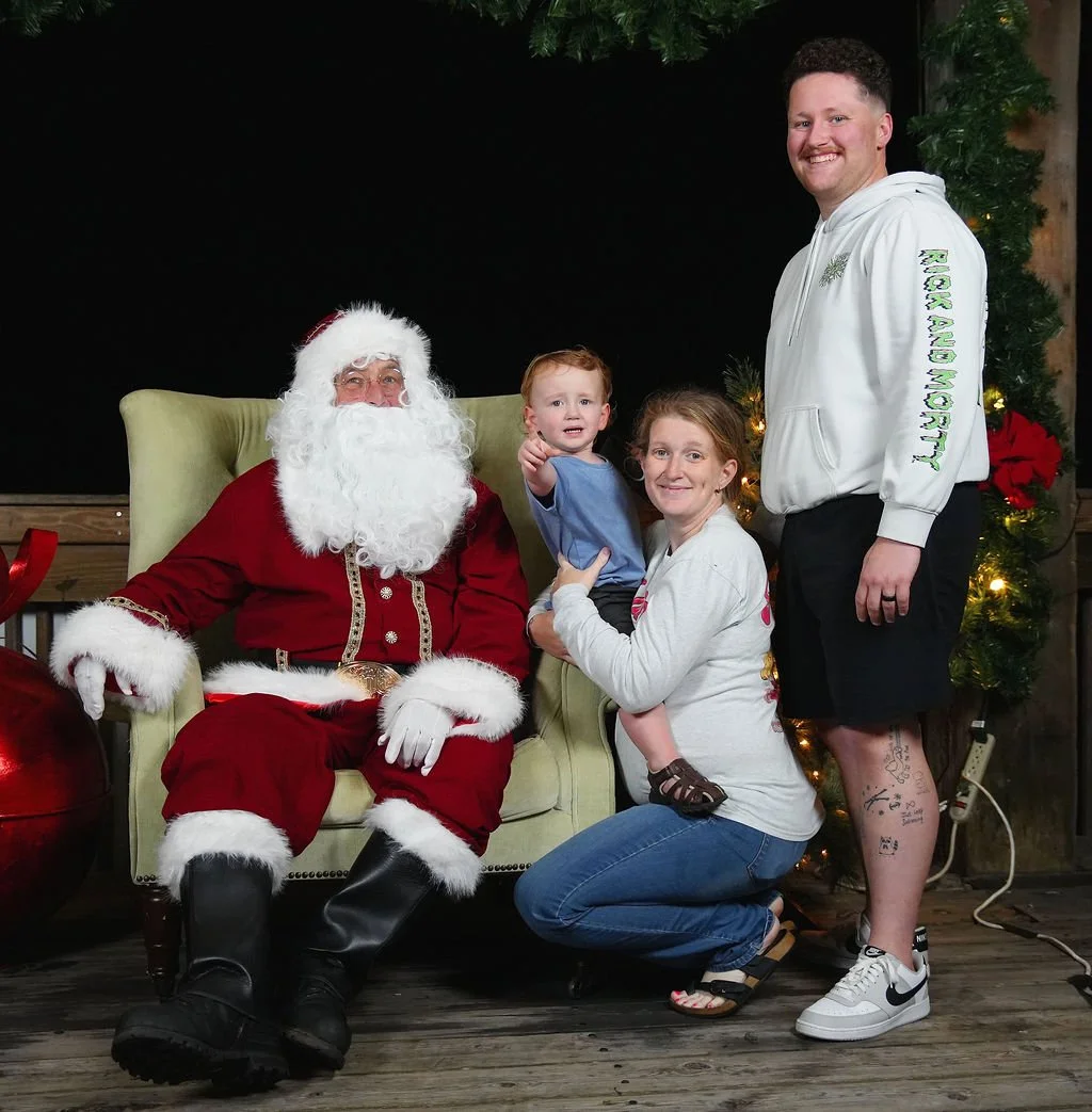 A man dressed as Santa Claus sitting on a green chair with a woman, a young boy, and a young man standing next to him. The woman is kneeling and holding the boy, who appears to be crying. The young man is smiling and standing to the right. There is a