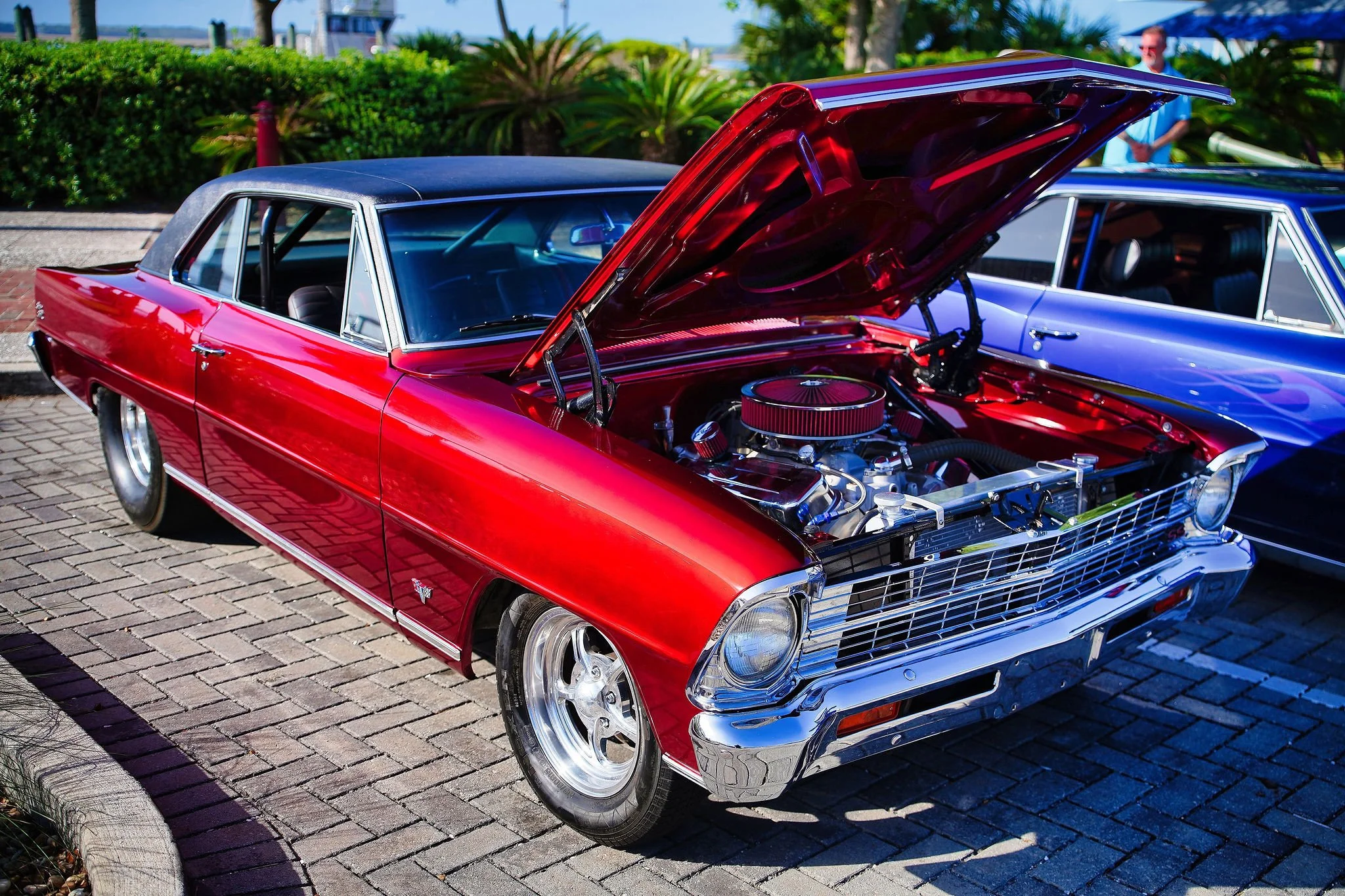 A red vintage car with its hood open at a car show, parked on a brick pavement near greenery.