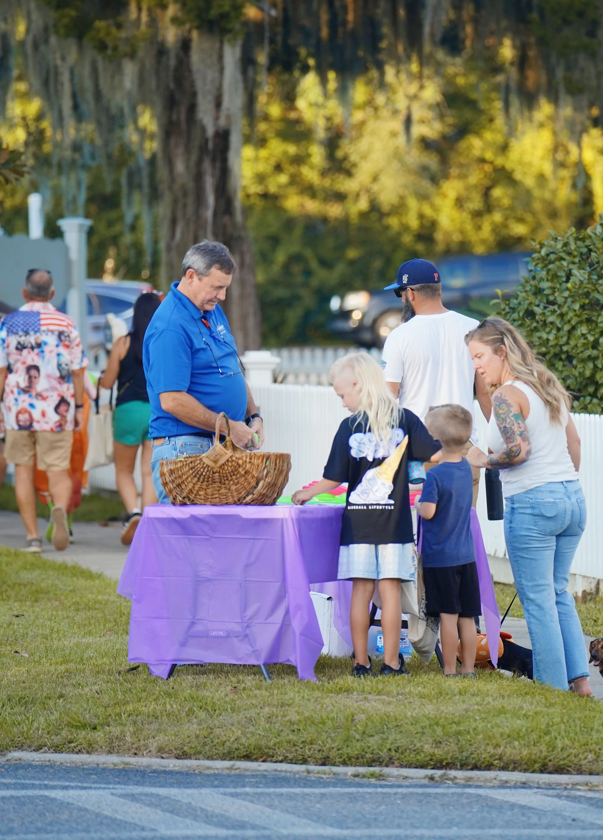A group of people gathered around a table covered with a purple tablecloth, outdoors in a park-like setting with trees in the background. The people are engaged in an activity, with some children and adults present.
