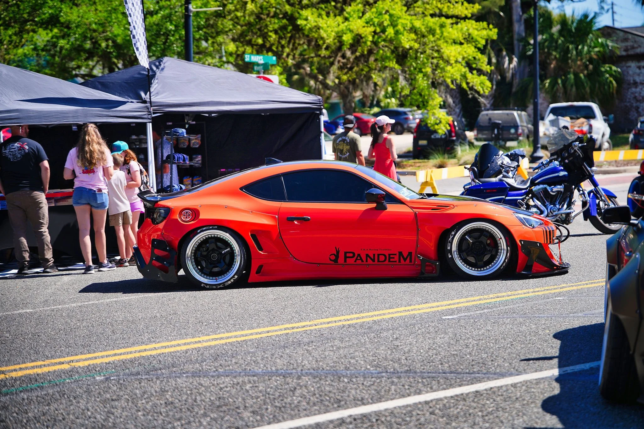 A red racing sports car with black wheels and a low profile parked on a street during a car event. There are several people and motorcycles nearby, and a black canopy tent with merchandise on displayed table behind the car. Green trees are in the bac