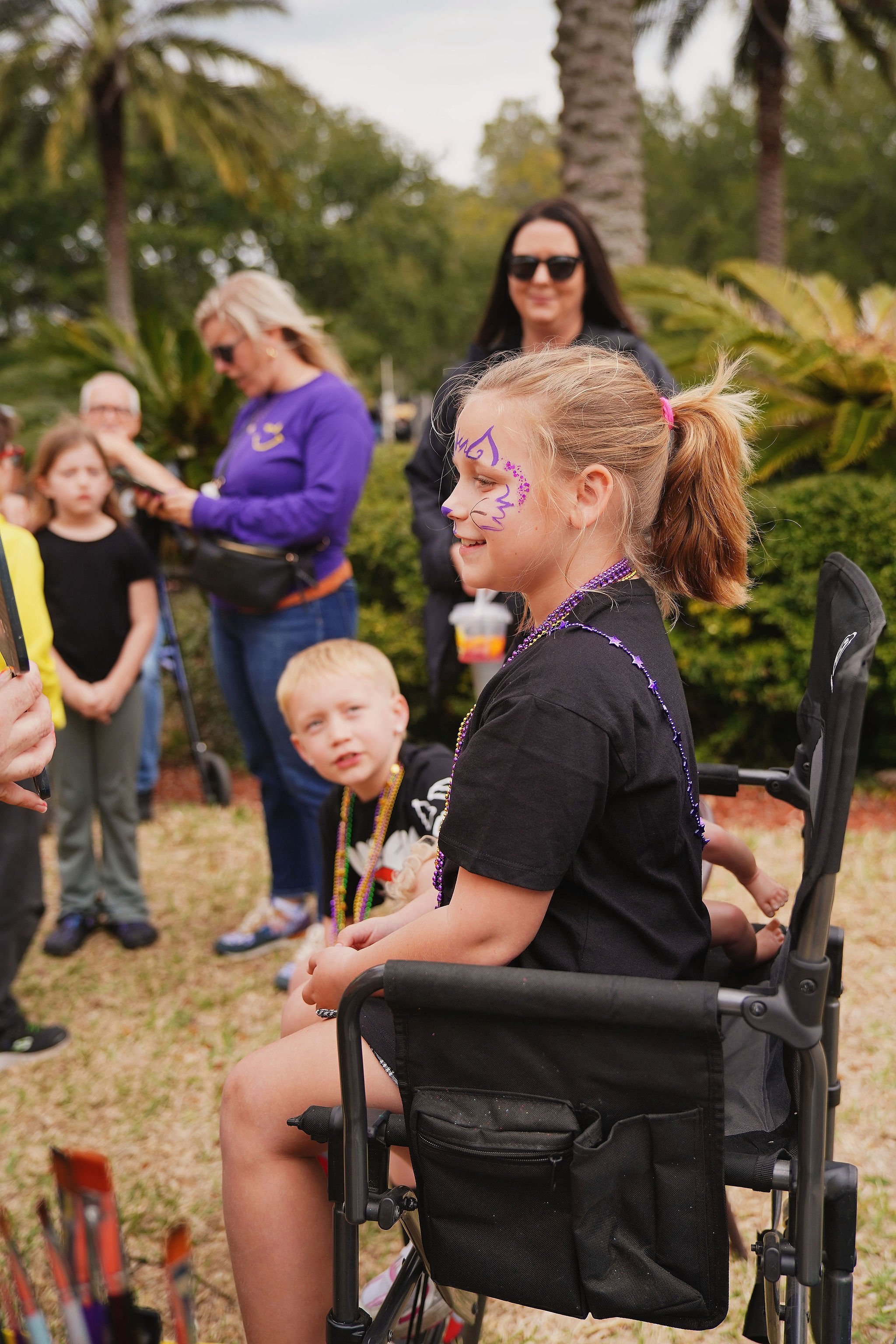 A young girl with face paint and beads sitting in a wheelchair at an outdoor event, with other children and adults around, some with face paint and wearing beads.