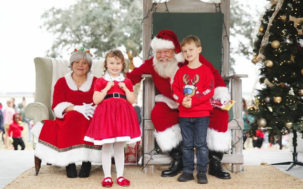 Children and Santa Claus sitting on a wooden throne next to a Christmas tree, with an older woman dressed as Mrs. Claus sitting nearby, all smiling for a holiday photo.