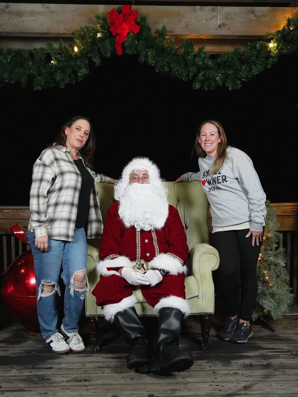 Two women standing next to Santa Claus, who is sitting in a chair, with Christmas decorations behind them, including a wreath with a red bow and a Christmas tree, on a wooden floor.