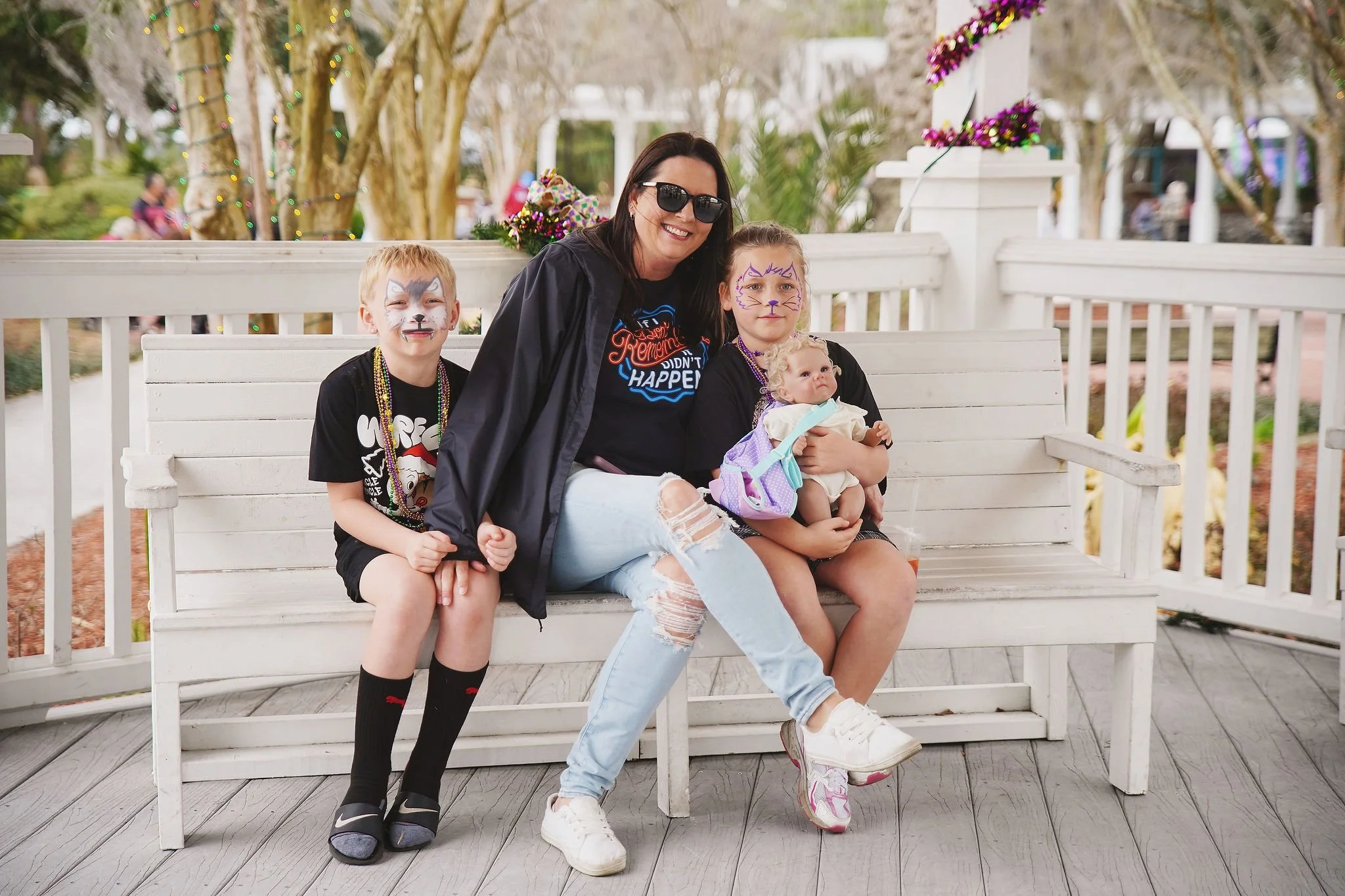 A woman with two children and a baby sitting on a white bench outdoors, with festive decorations and trees in the background.