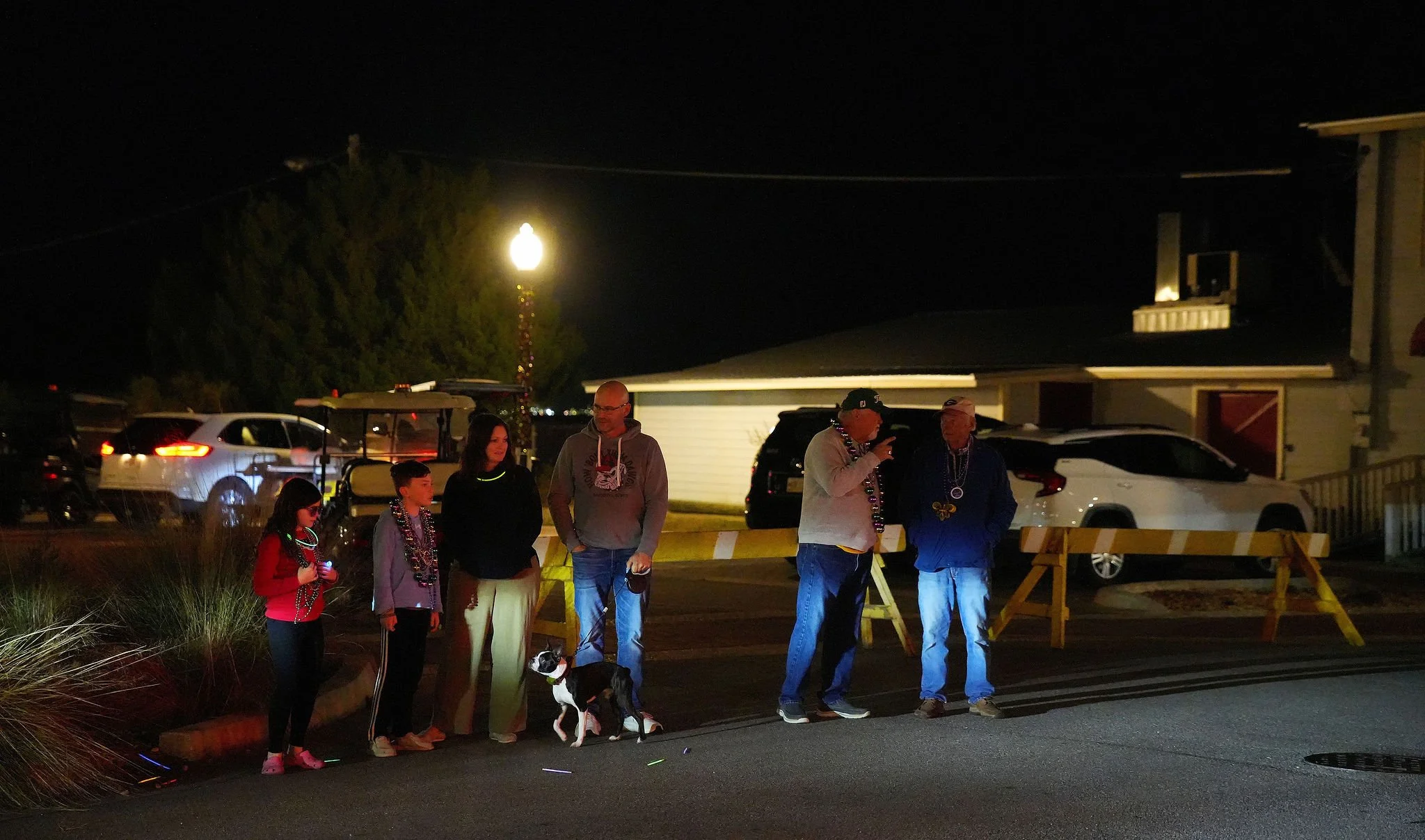 Group of six people, including children, standing outside at night decorated with beads and necklaces, with one person holding a dog, near a barricade, cars parked in the background, and a lit streetlamp.