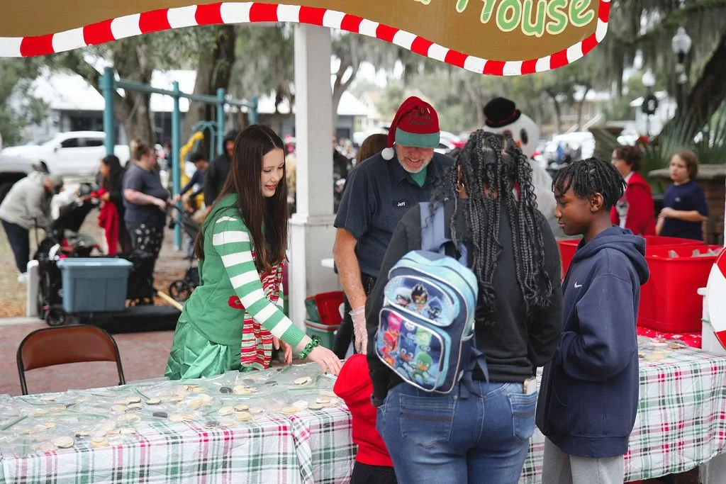 People at a holiday gift exchange or fundraiser event outdoors, with a table full of cookies and festive decorations.