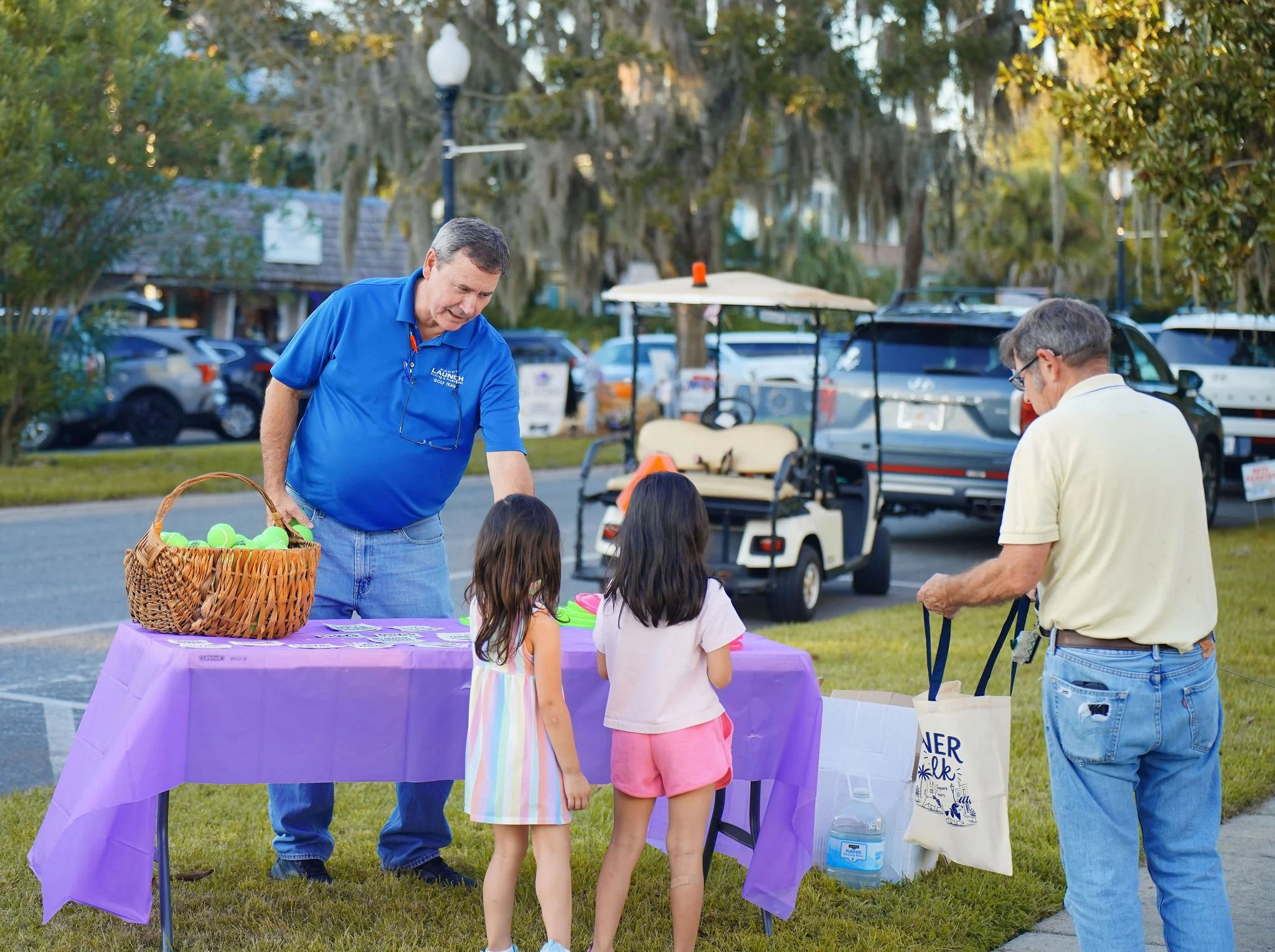 People gathered at a community event with a table covered in a purple tablecloth, a basket of tennis balls, and informational materials. Two men are engaging with children near the table. The background features parked cars, trees with hanging moss, 