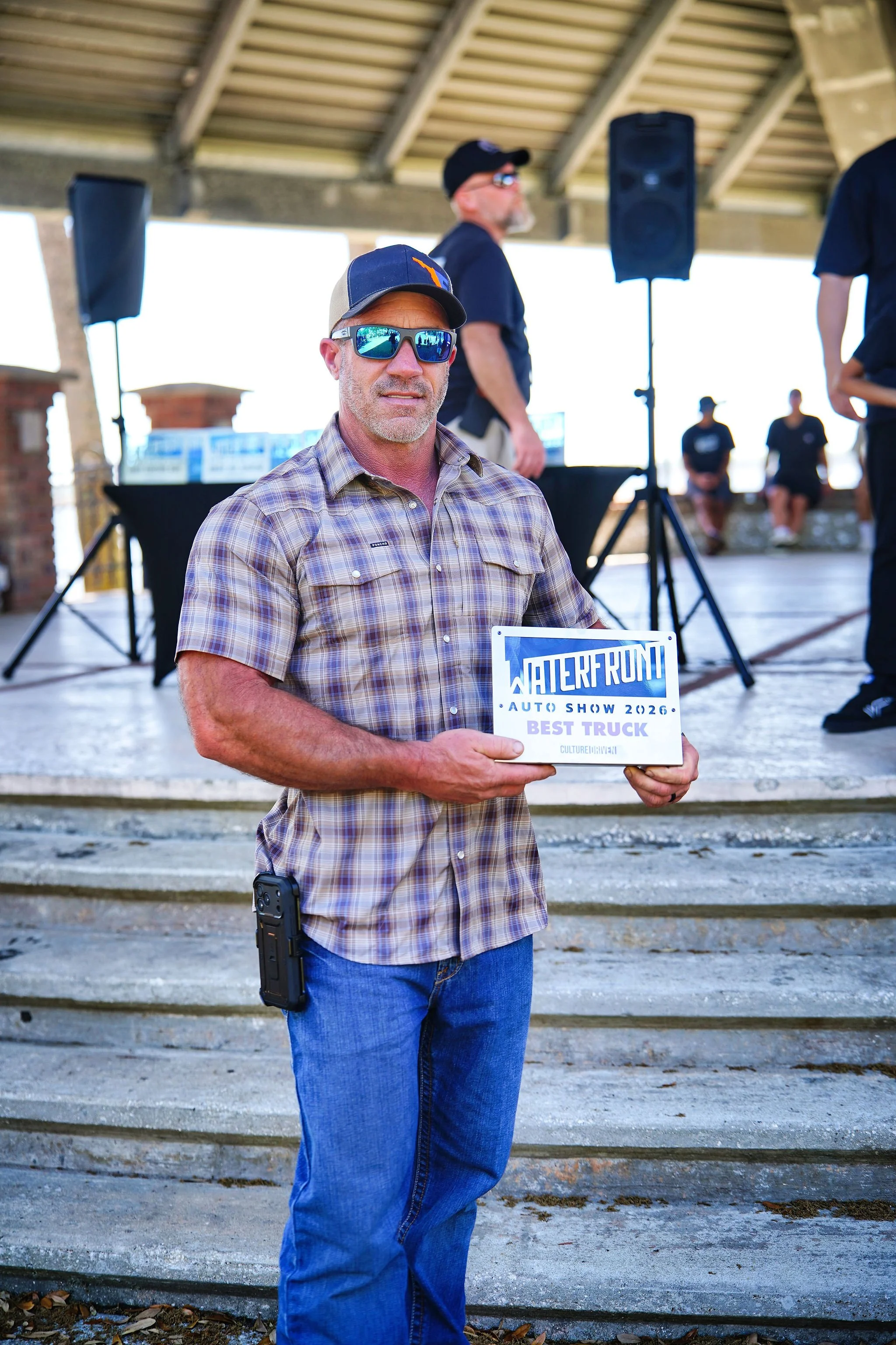 A man wearing sunglasses and a hat holds a sign that reads 'Waterfowl Auto Show 2026 Best Truck' at an outdoor event, with a stage and other people in the background.
