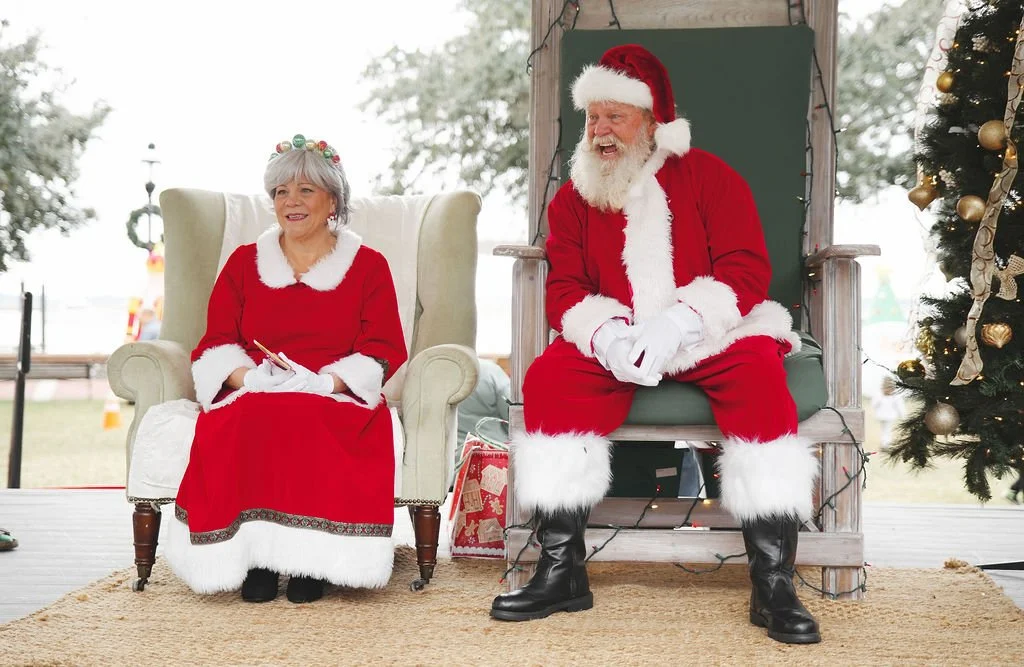 A woman and a man dressed as Santa Claus sitting on stage during a Christmas event. The woman is seated in a white armchair, wearing a red dress with white trim, and the man is seated in a wooden throne, wearing a red Santa suit with black boots and 