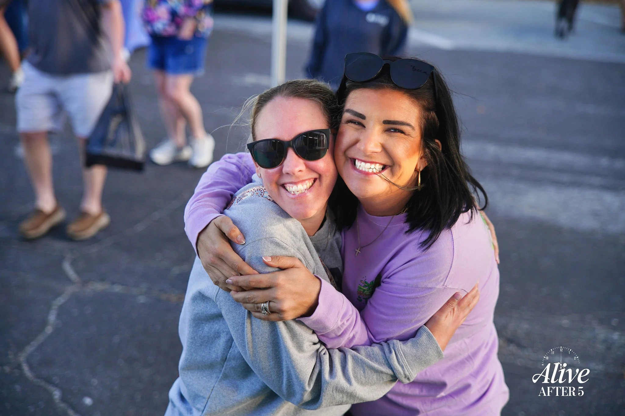 Two women hugging and smiling at the camera, outdoors, with other people in the background, one wearing sunglasses and a grey hoodie, the other wearing a lavender shirt and sunglasses on her head.