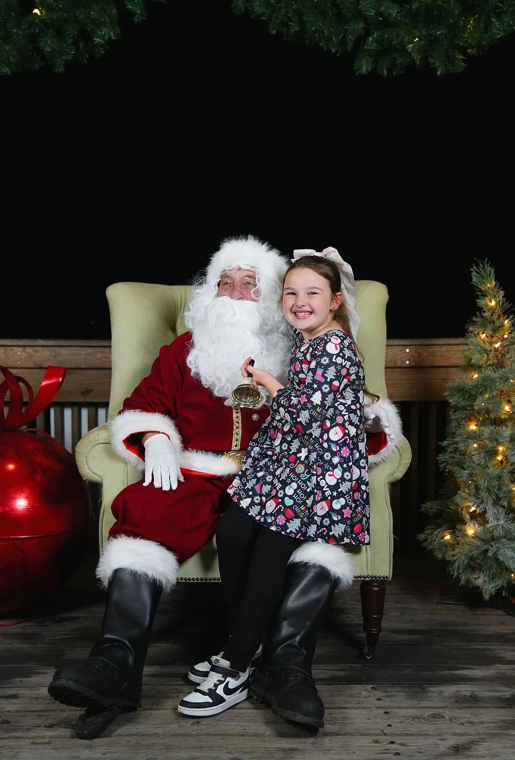 A young girl sitting on Santa Claus's lap, holding a small bell, smiling. Santa is seated in a green armchair, wearing a red suit with white fur trim, and has a white beard and glasses. Christmas decorations, including a Christmas tree decorated with