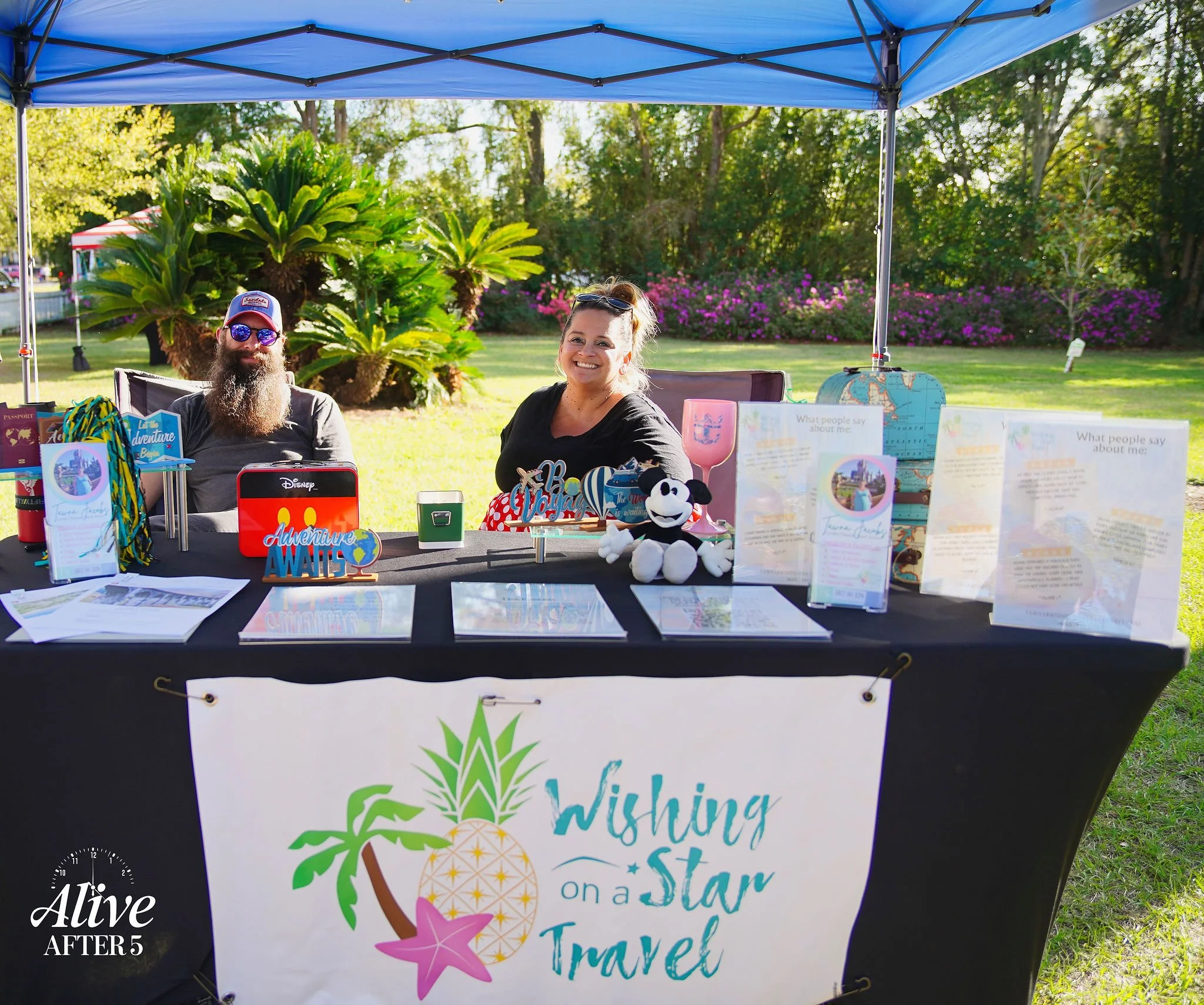 Two smiling people sitting behind a table with travel-themed items under a blue canopy in a park, with trees and flowers in the background. The table displays brochures, a Disney adventure suitcase, a globe, a Mickey mouse plush, and a banner that re