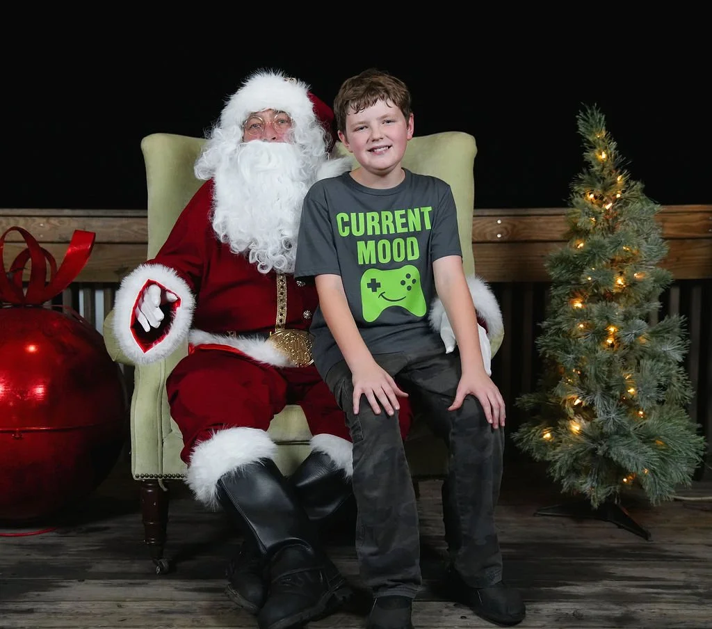 A boy sitting on Santa Claus's lap during a Christmas photo, with a decorated Christmas tree and a large red ornament in the background.