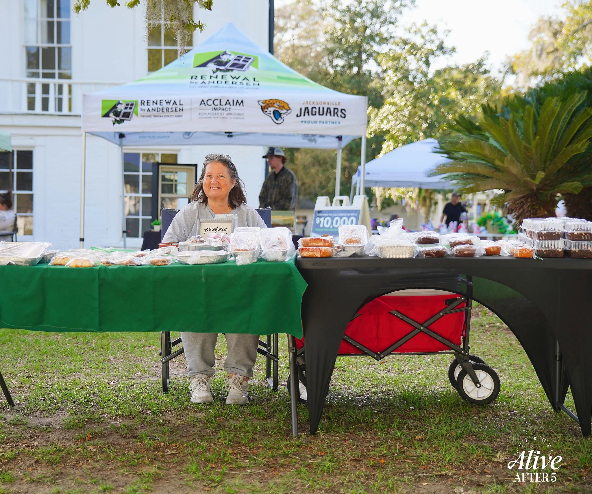 Smiling woman standing behind a table with packaged baked goods at an outdoor event, under a white canopy with Jacksonville Jaguars and other logos, on grass with trees and other tents in the background.