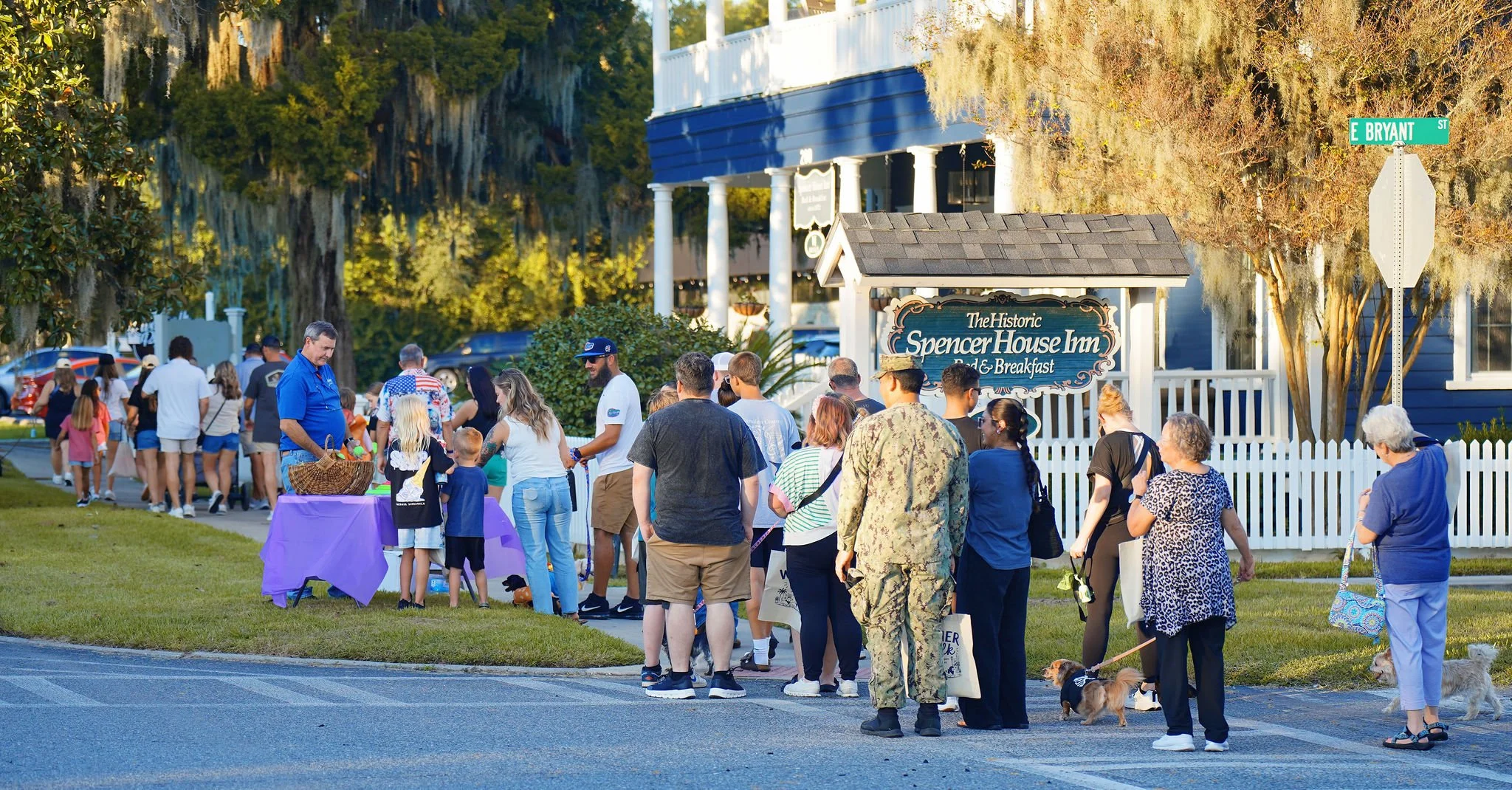 A group of people waiting in line outside Spencer House Inn Bed & Breakfast on a sunny day, with some people walking dogs and others chatting, near a street sign for E Bryan Street.