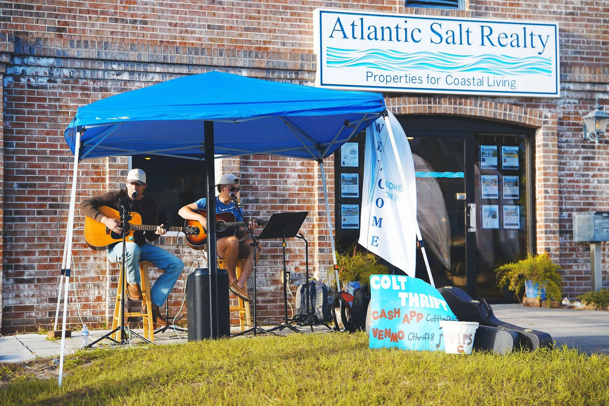 Two young men perform acoustic guitar music under a blue canopy outside a building with a sign for Atlantic Salt Realty. There are signs promoting a thrift store and digital sales, with backpacks and guitar cases on the ground.