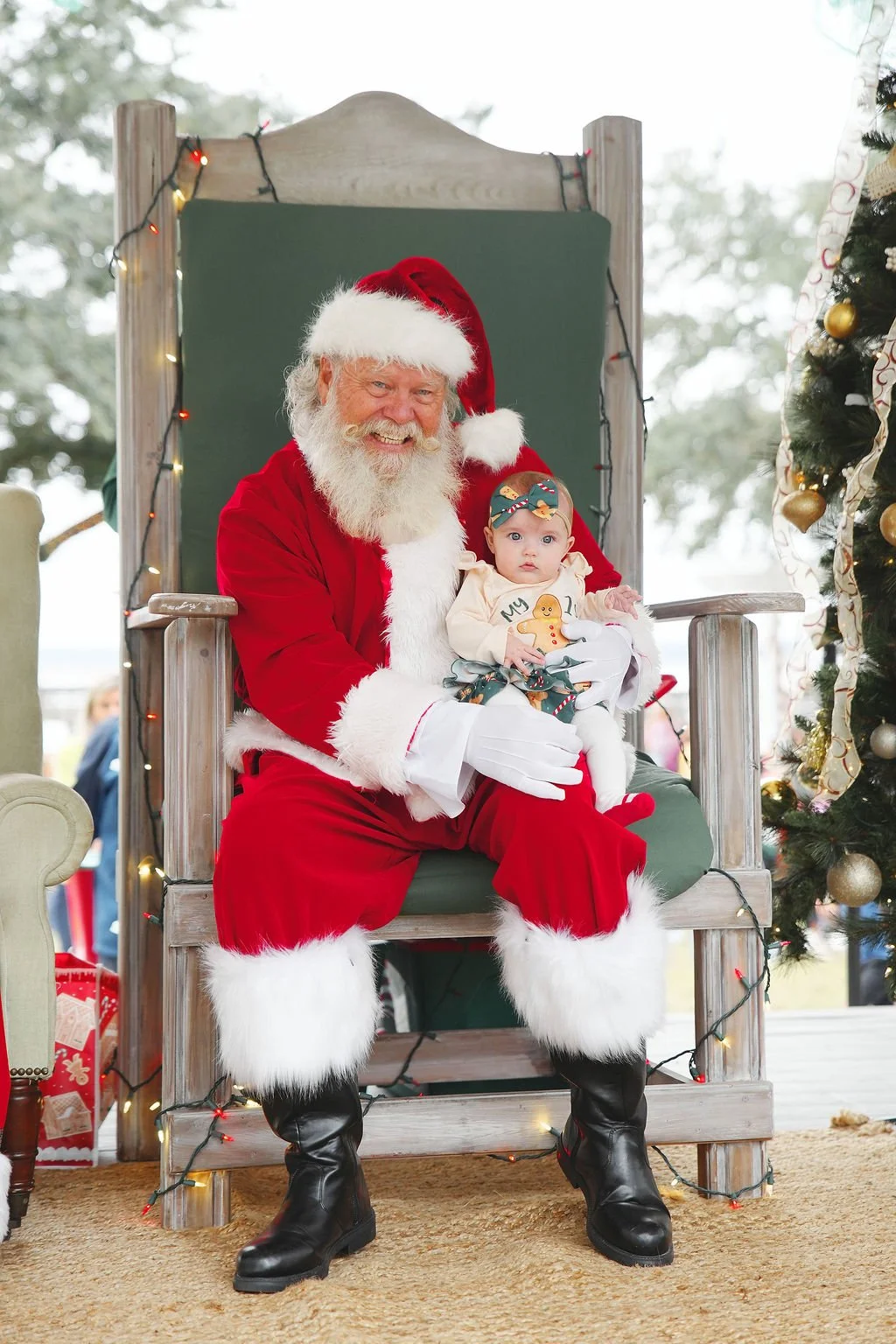 Santa Claus sitting on a large wooden chair holding a baby girl dressed in holiday attire, with Christmas decorations and a Christmas tree nearby.