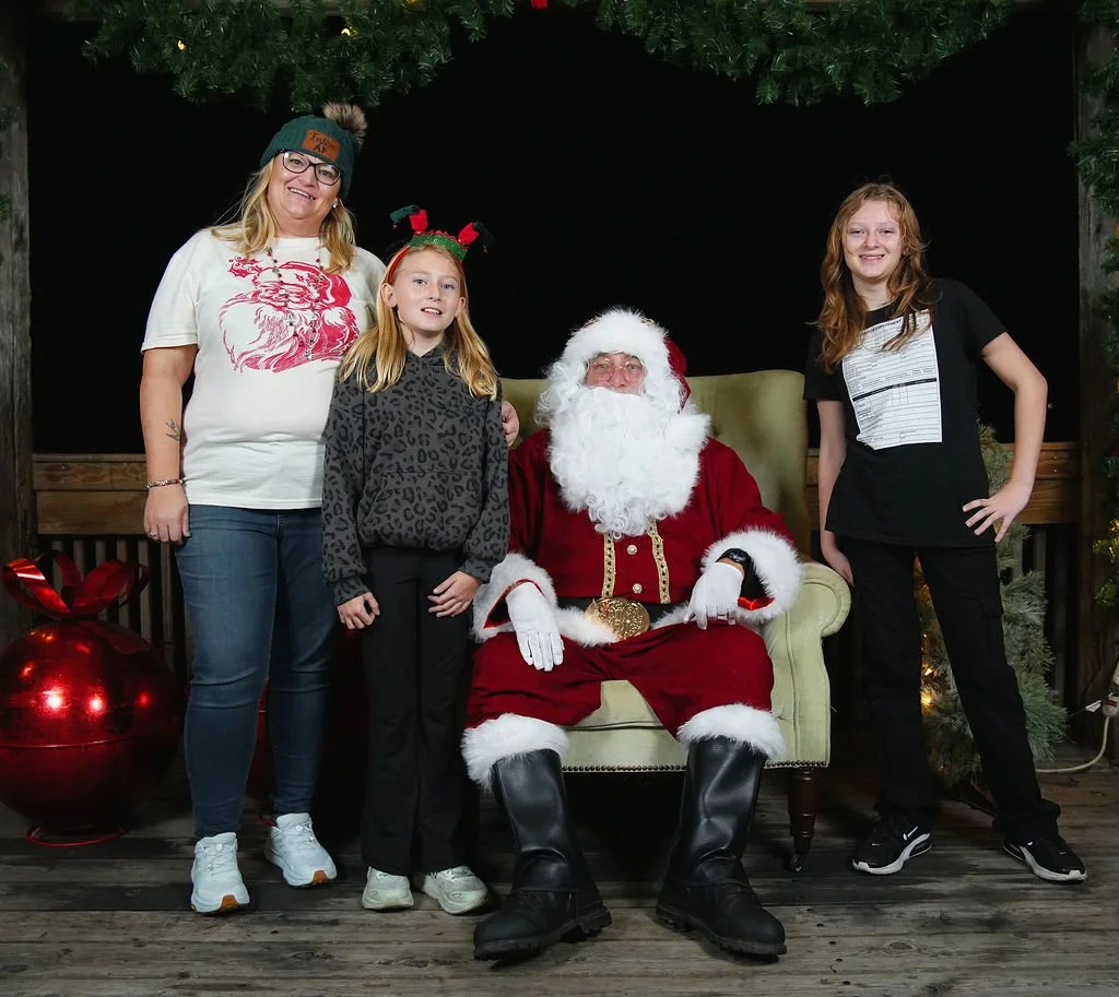 Four women and Santa Claus sit and stand in front of a Christmas backdrop. Santa is seated on a green armchair, wearing his traditional red suit, black boots, and a white beard. The women are dressed casually, with one wearing a white t-shirt with a 