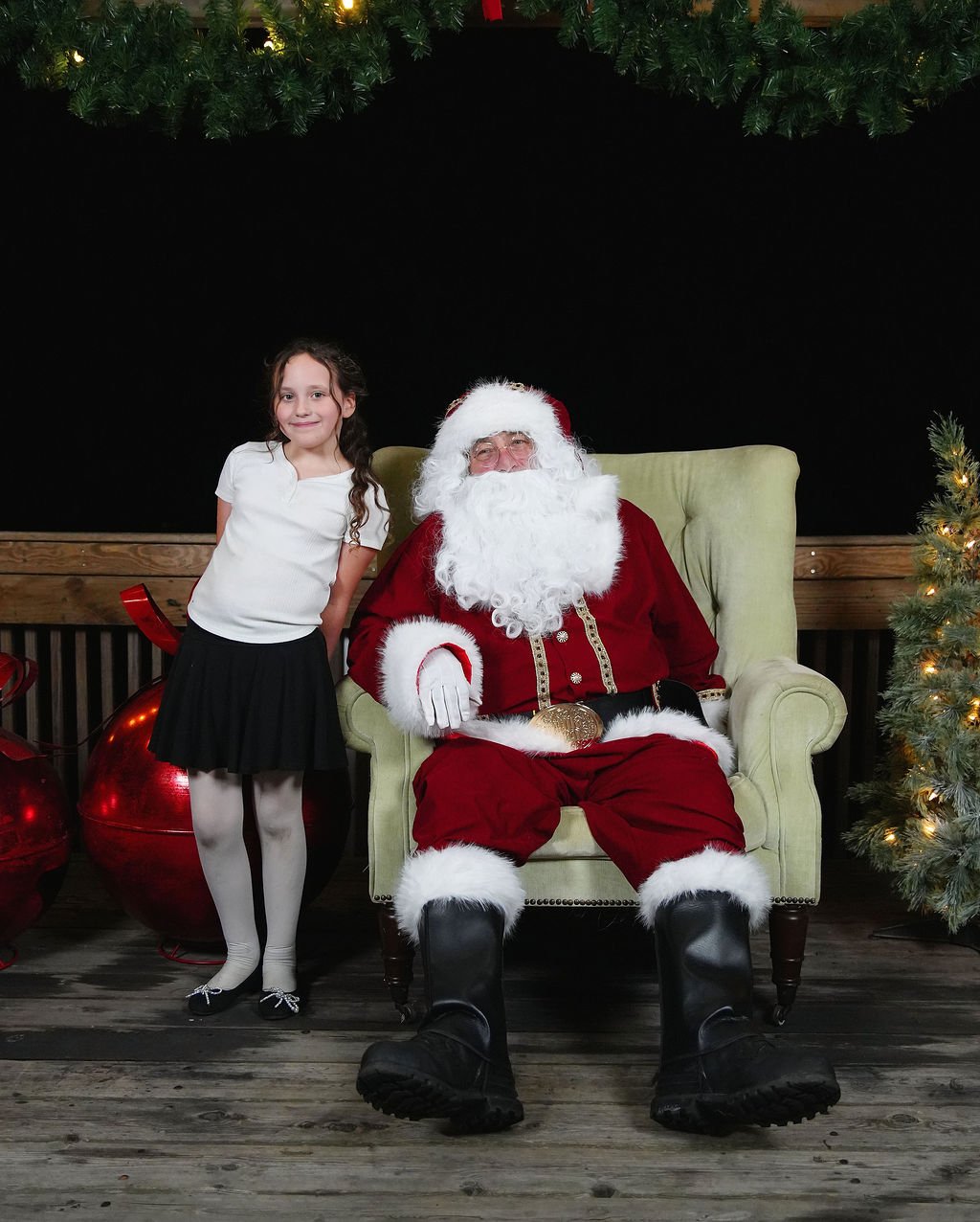 A young girl with long curly hair, wearing a white blouse, black skirt, white tights, and black shoes, standing next to Santa Claus seated in a green armchair. Santa has a white beard, red suit, black boots, and a gold belt. The background has Christ