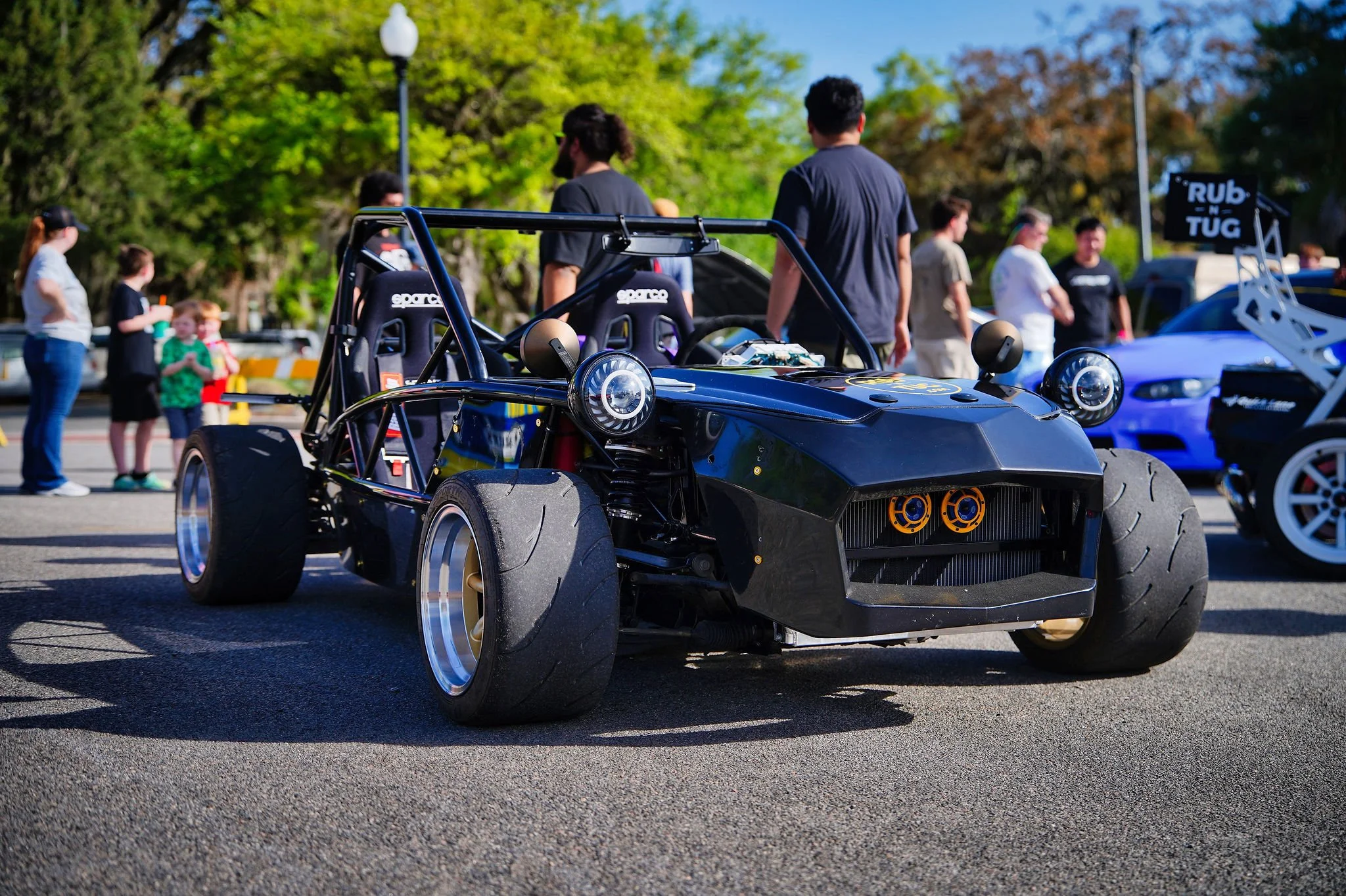 A black race car with wide tires and an exposed chassis parked on a street during an outdoor car show, with people walking and looking at other cars in the background.