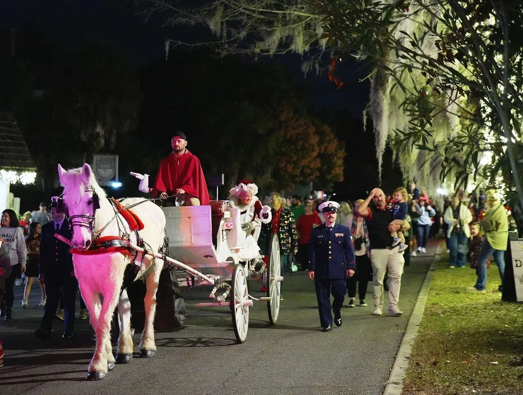 A nighttime scene at a holiday parade featuring a horse-drawn carriage with actors dressed as Santa and Mrs. Claus, people walking along the float and sidewalk, some in festive attire, and trees lit with decorations.