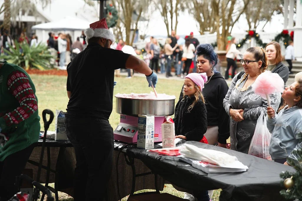Person wearing a Santa hat making cotton candy at an outdoor holiday event, with people waiting in line and decorations in the background.