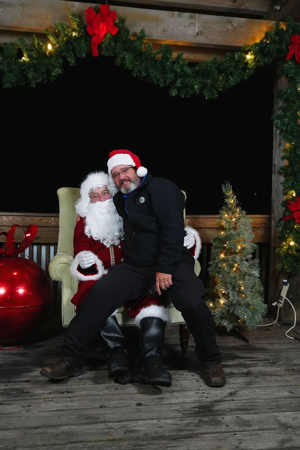 A person dressed as Santa Claus sitting on a large green chair with a person in black sitting on his lap, both smiling. The scene is decorated with green garlands, red ribbons, a small Christmas tree with lights, and a large red ornament, in a rustic