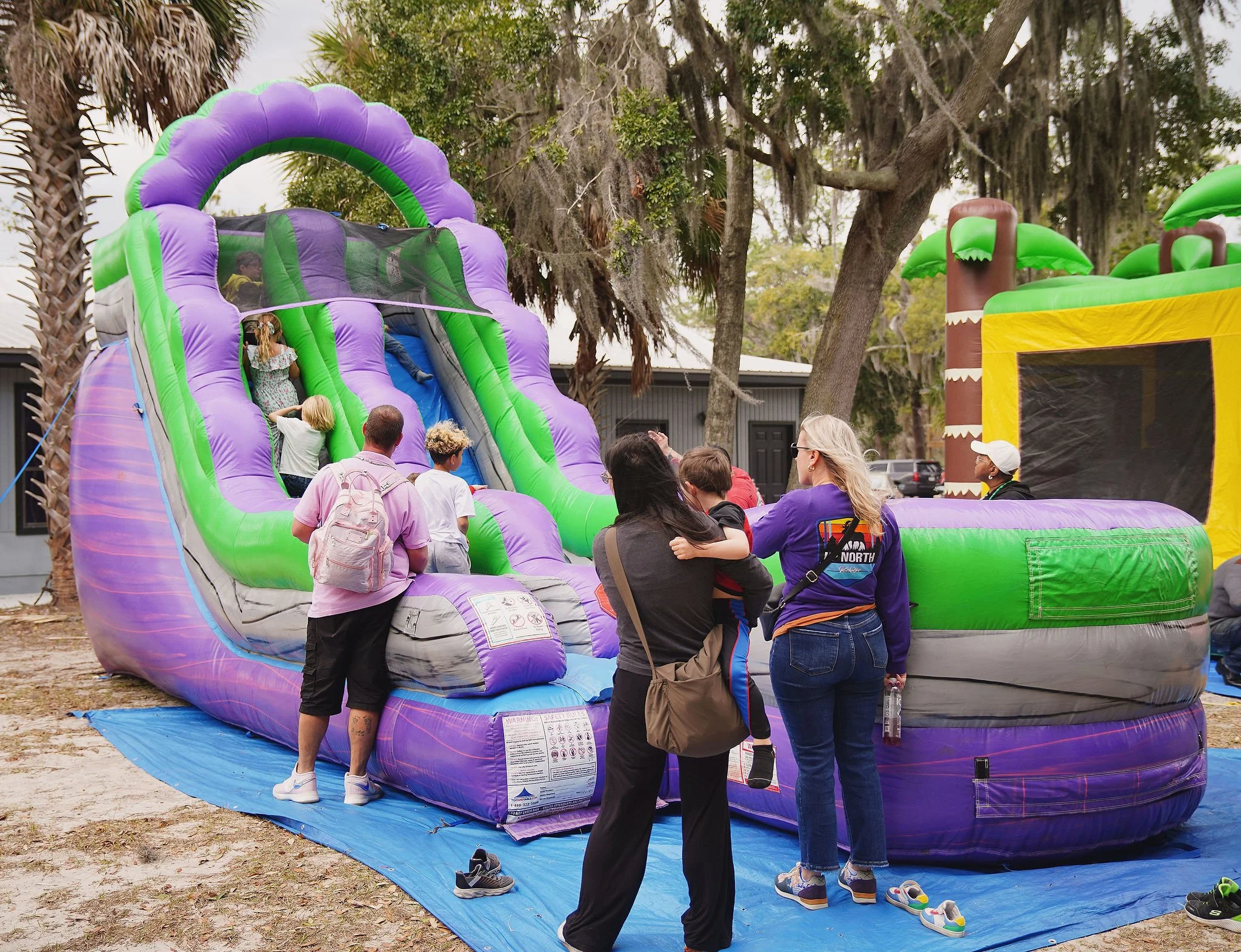 Children playing on an inflatable water slide at an outdoor amusement park, with adults supervising and watching nearby.