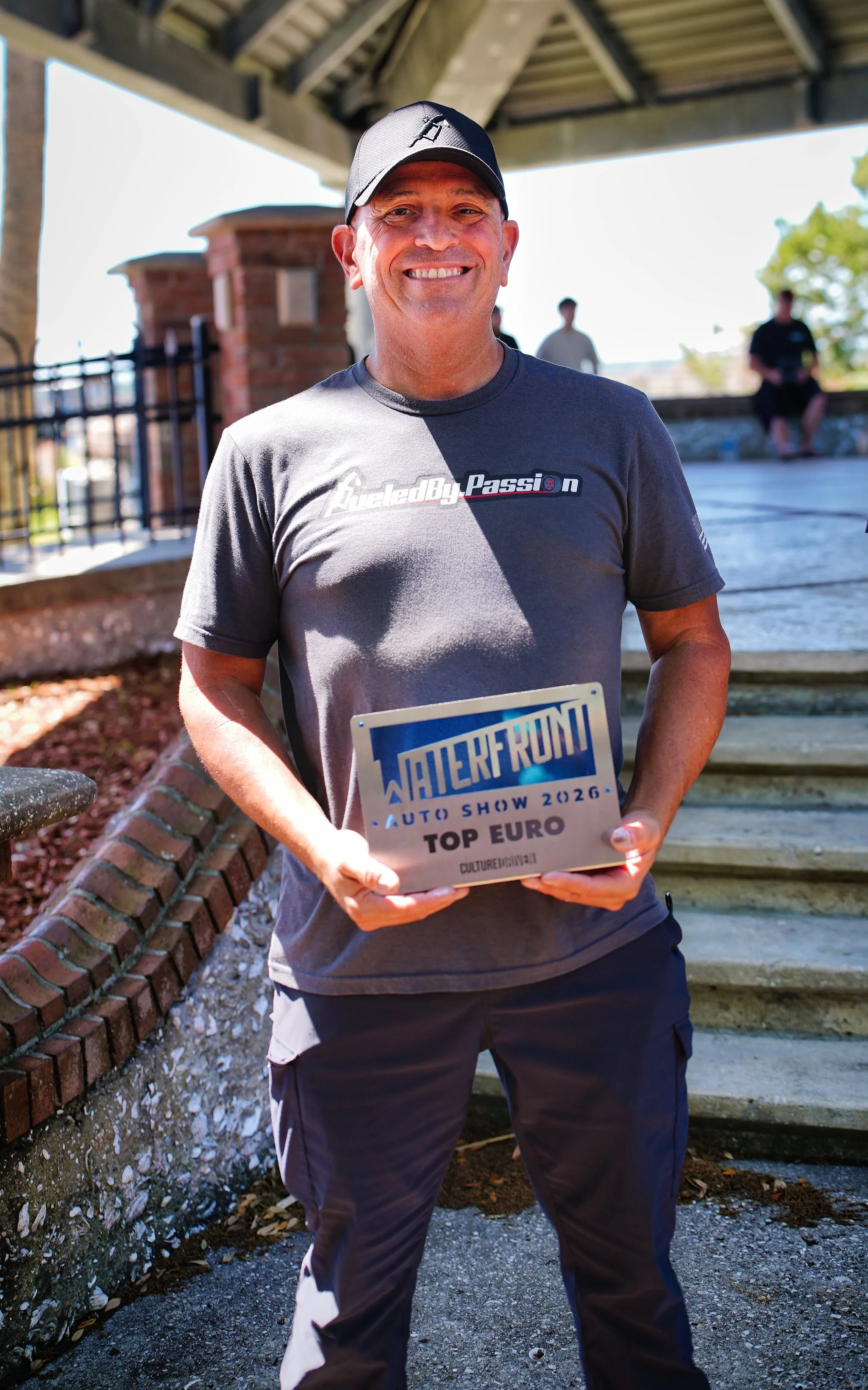 A man smiling while holding a trophy at an outdoor event, wearing a black cap and a gray T-shirt with 'Fueled by Passion' written on it. The trophy reads 'WaterfRUND Auto Show 2026 Top Euro' and the background shows a staircase and two other people s