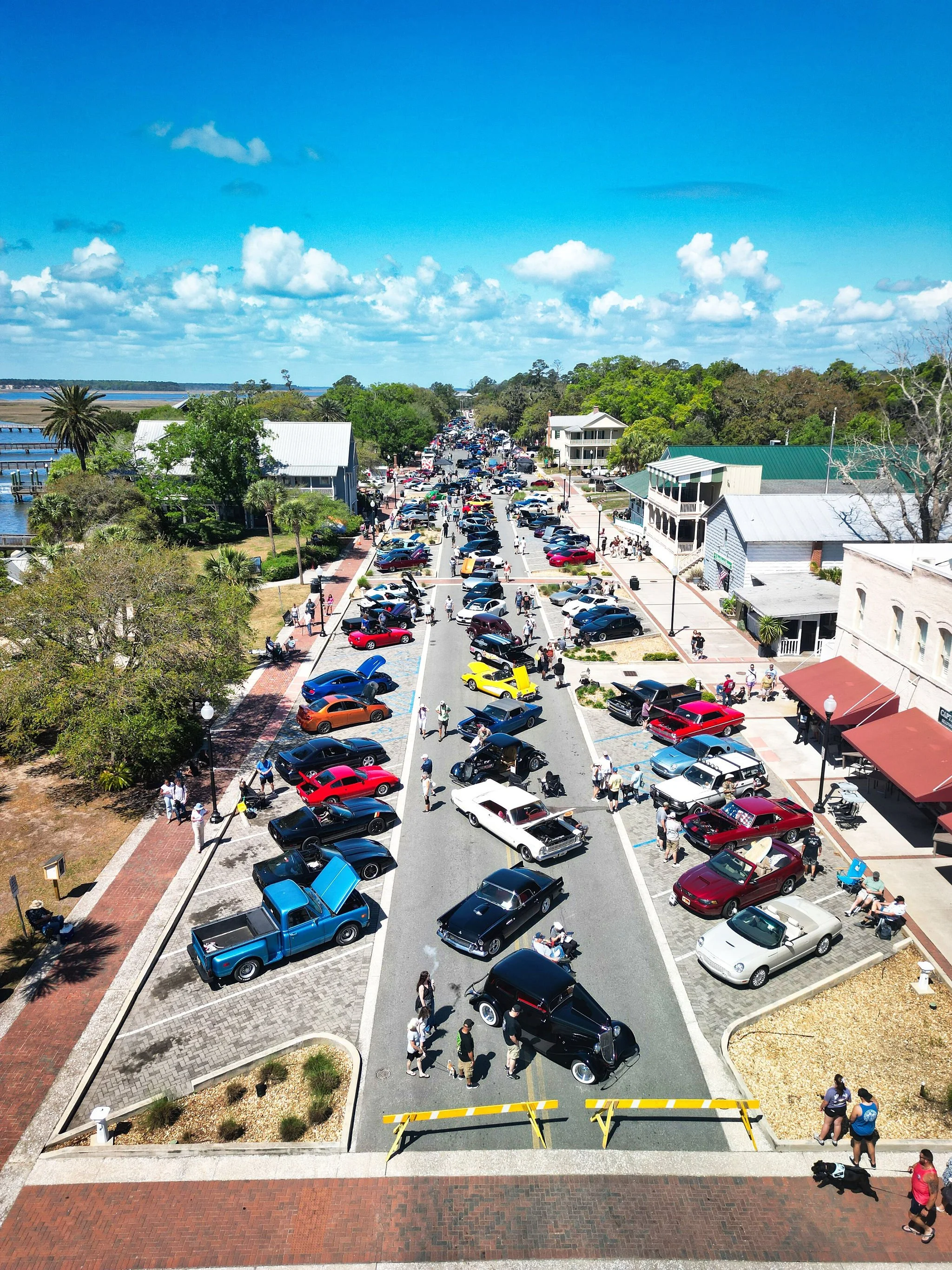 Aerial view of a busy street lined with classic cars and people, with buildings on either side, green trees, and a blue sky with clouds.