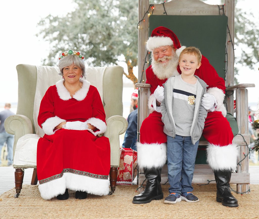 A young boy sitting on Santa Claus's lap with Mrs. Claus sitting nearby during a holiday event.