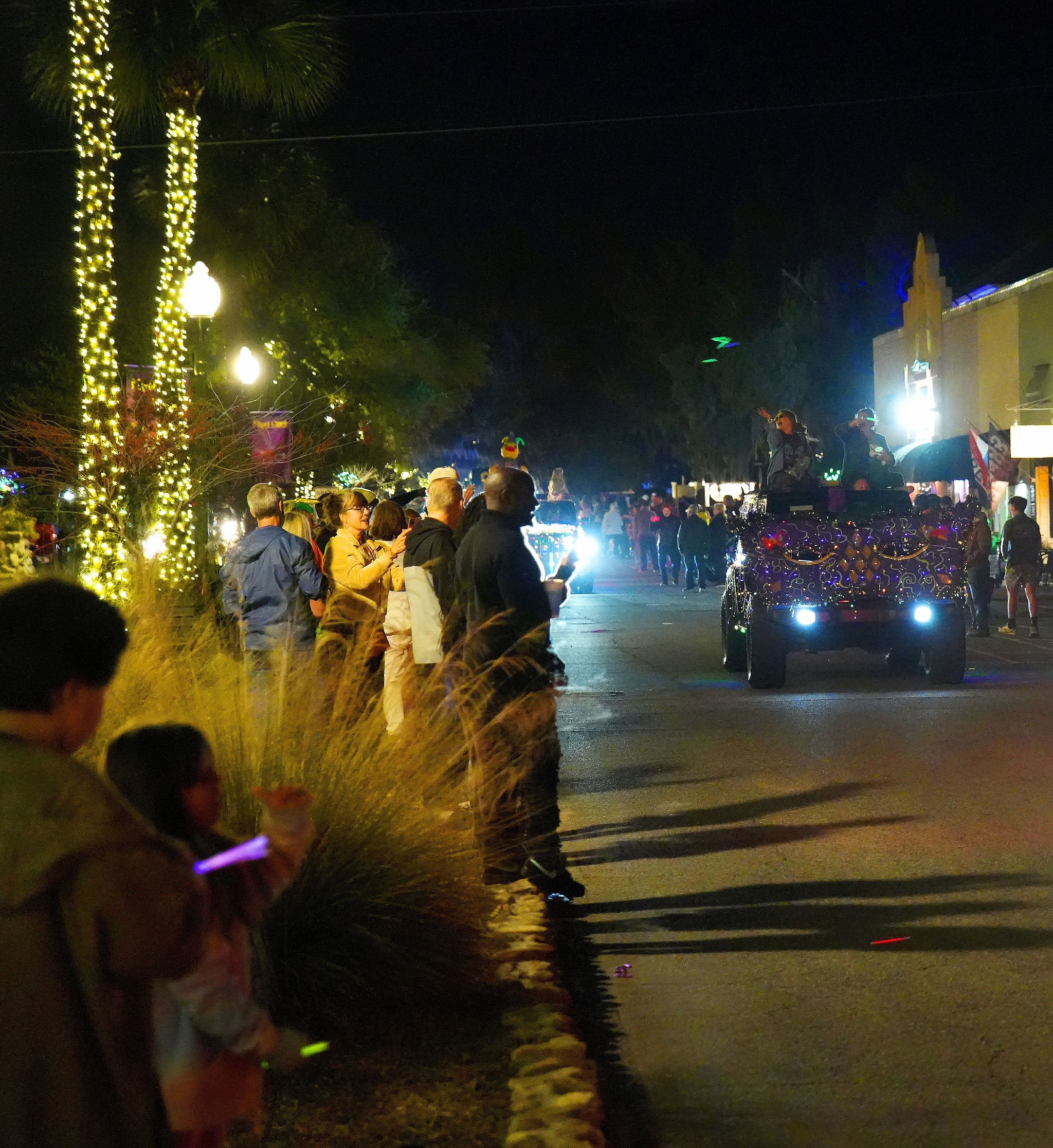 A nighttime parade with people lined up along the street watching decorated floats pass by, with lights and festive decorations, in a small town.