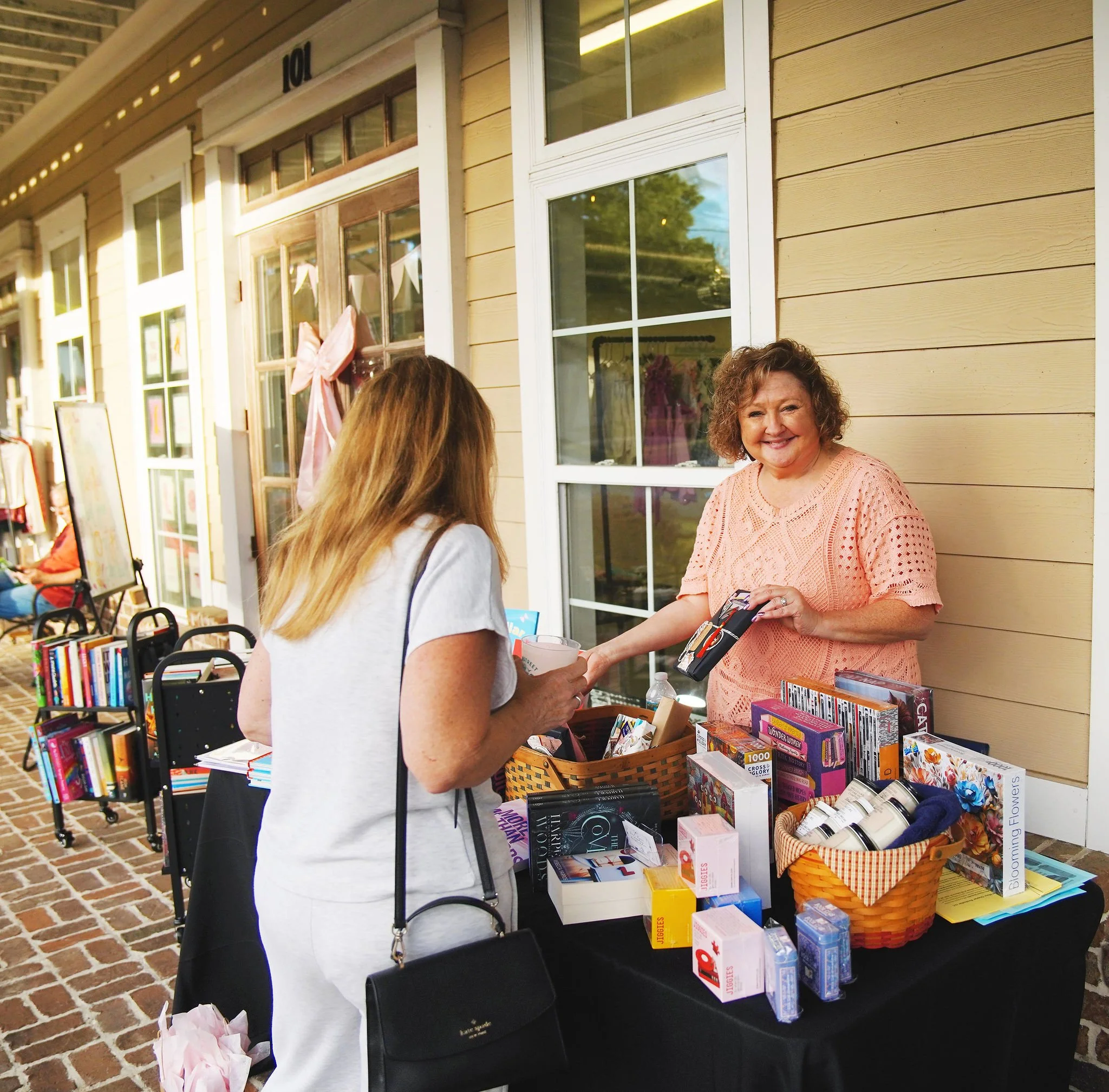 A woman with curly hair and a peach-colored top sells items at an outdoor table, smiling, while another woman in a white shirt and purse shops nearby.