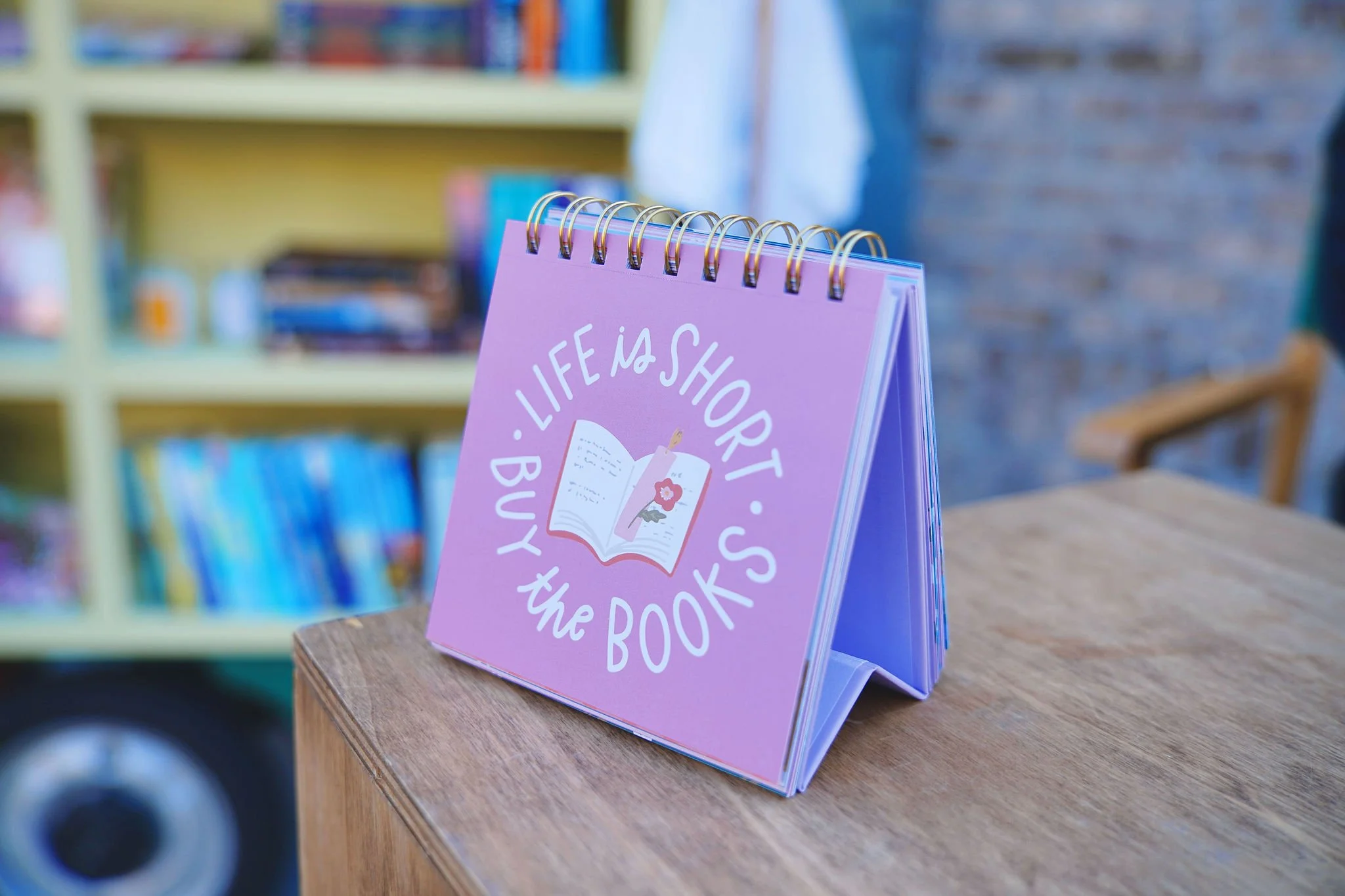 A pink desk calendar with the quote "Life is short, buy the books" and an illustration of an open book and a bookmark, placed on a wooden table in a cozy room with bookshelves in the background.