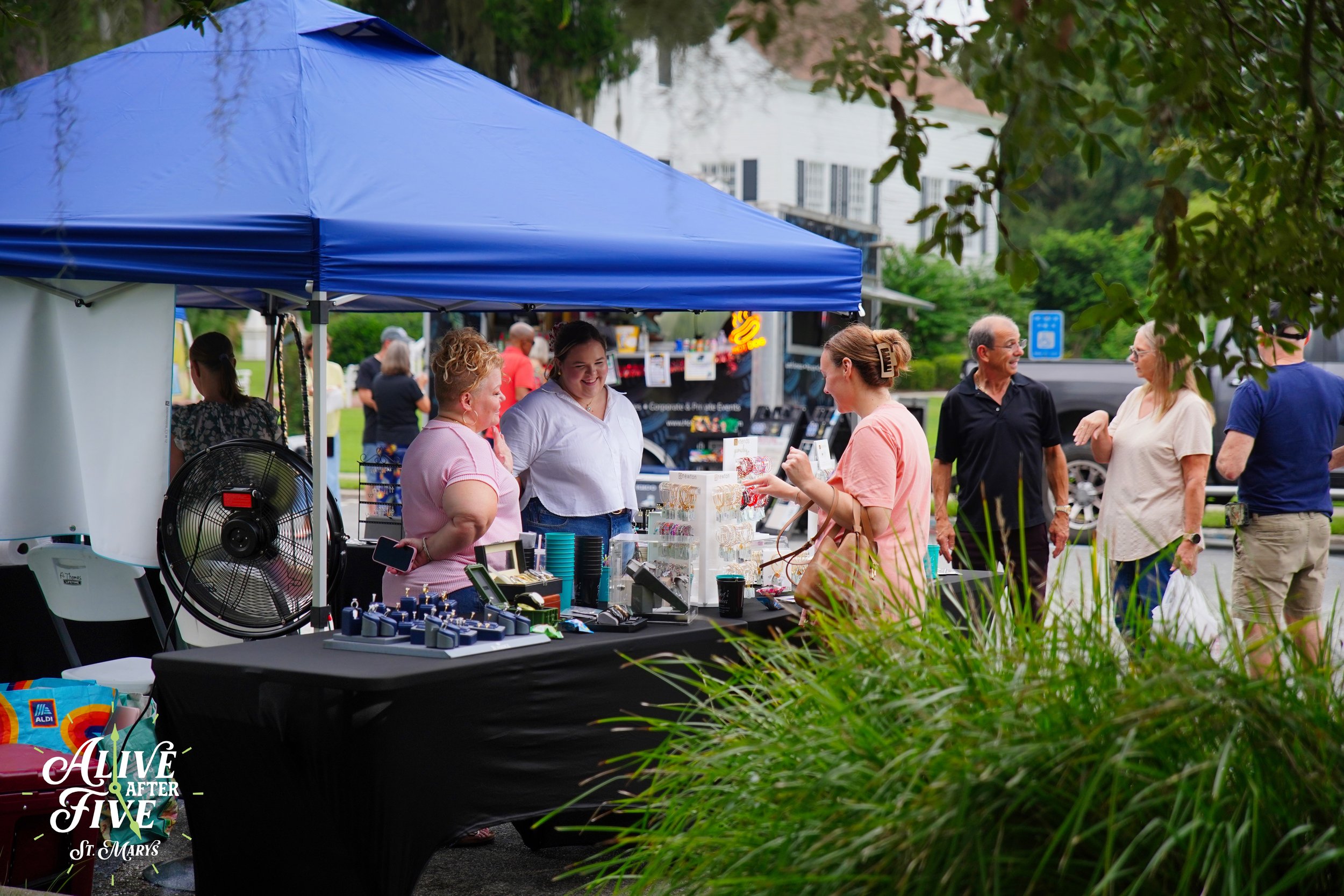 People shopping at an outdoor market stall under a blue canopy with jewelry and accessories on display. Several people are talking and browsing, with trees and a white building in the background.