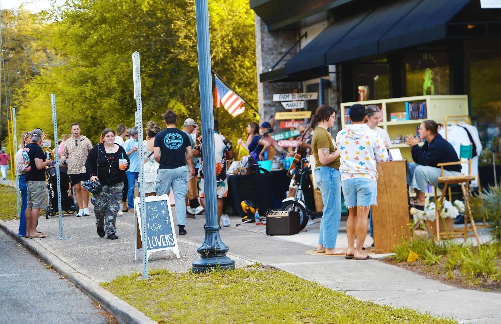 People shopping at an outdoor market on a sidewalk, with a sign reading 'Buy Your Wife Flowers,' and a few merchants and a musician performing on the street. The scene takes place in a green, tree-lined area in the late afternoon or early evening.