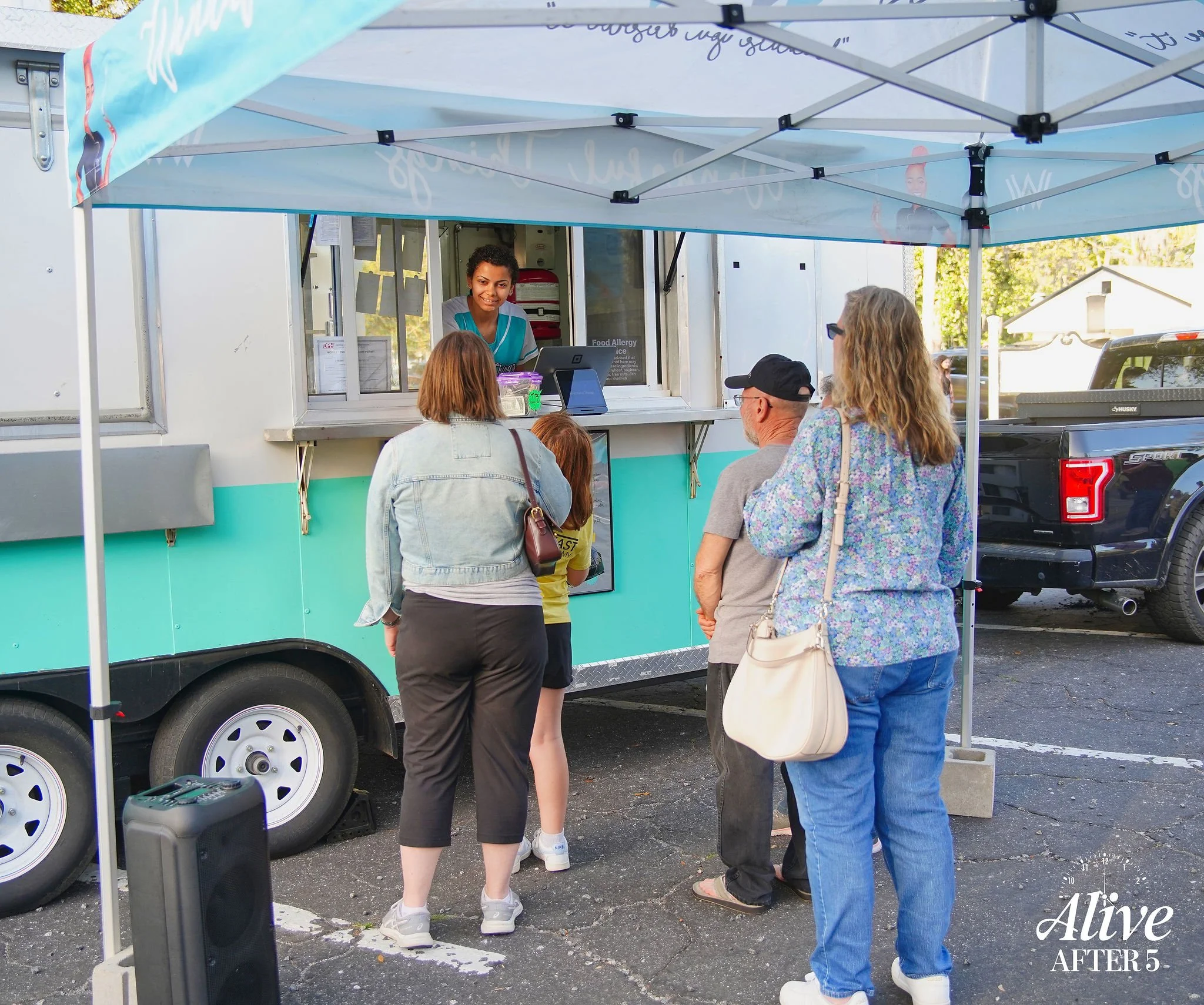 People ordering food at a food truck outside on a sunny day, with a woman woman serving through the window, underneath a blue canopy.