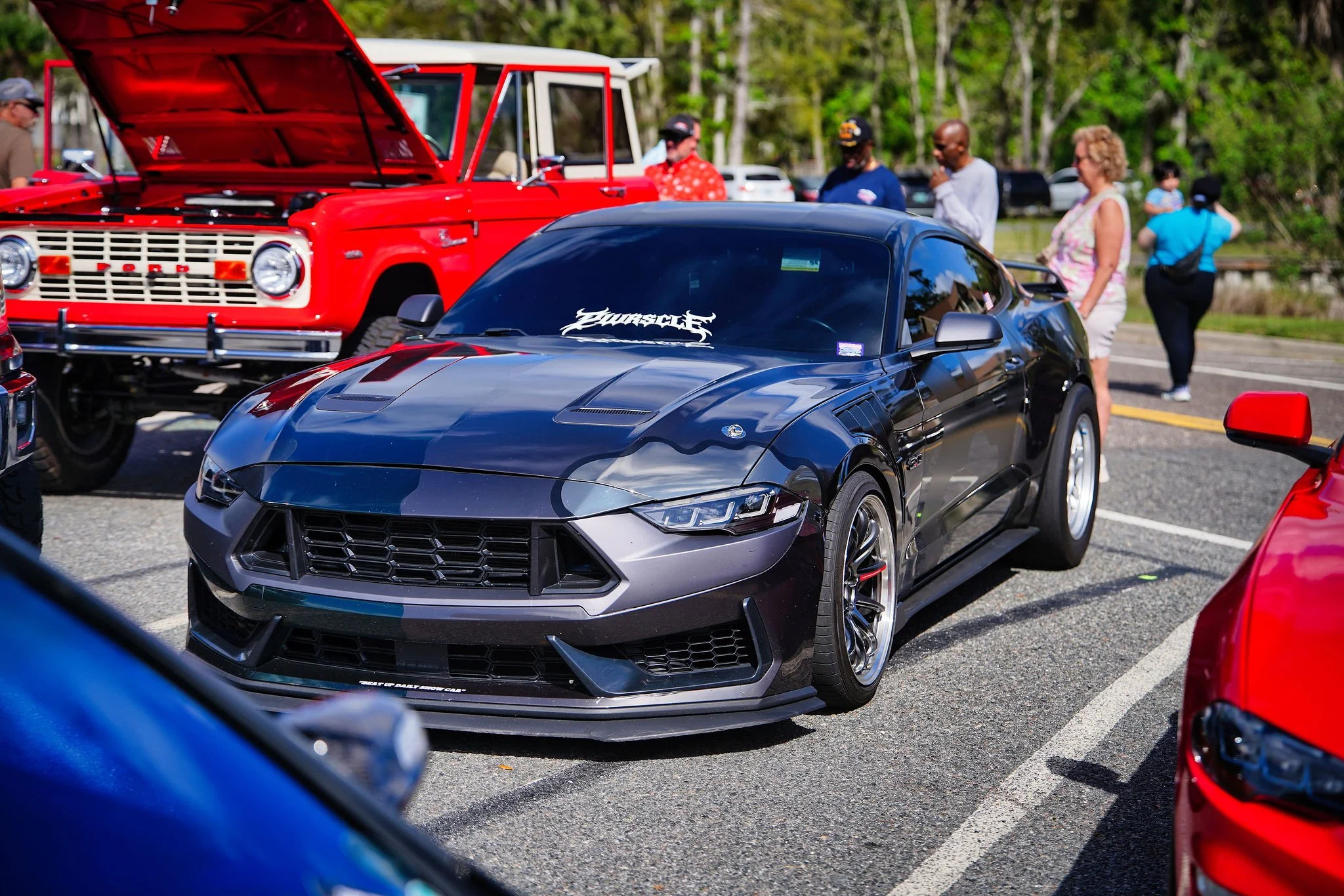 A black sports car parked among other vehicles at a car show, with people walking and looking at the cars in the background.