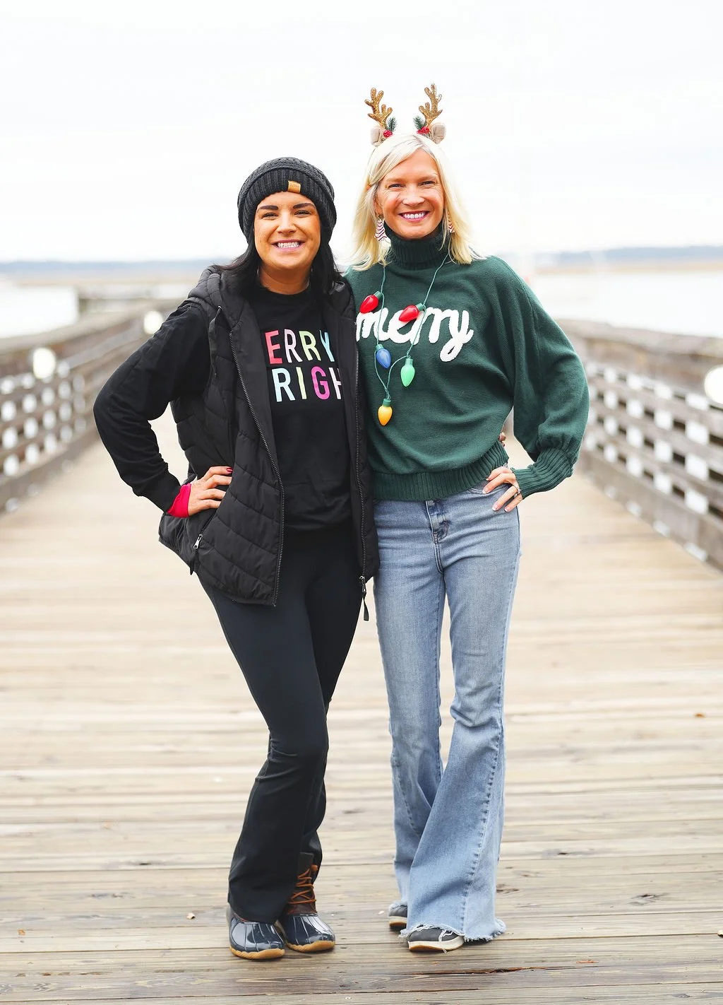 Two women standing on a wooden pier outdoors, smiling. One is wearing a black jacket, beanie, and a Christmas-themed shirt. The other wears a green sweater with 'Merry' and Christmas lights, and reindeer antlers headband.