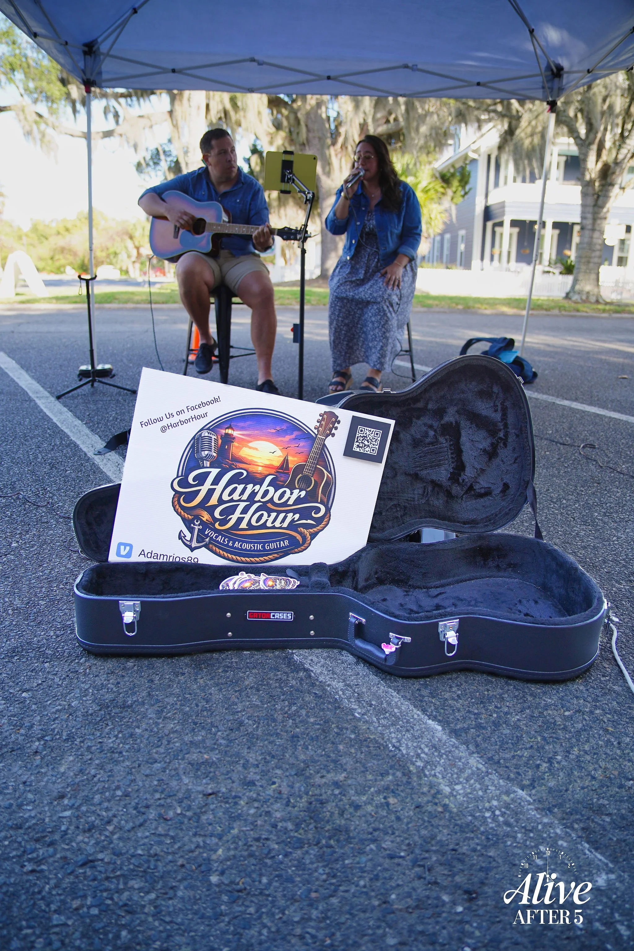 A street performer setup includes an open guitar case with a Harbor Hour sign, two musicians under a blue canopy, one playing guitar and the other singing, with a residential neighborhood in the background.