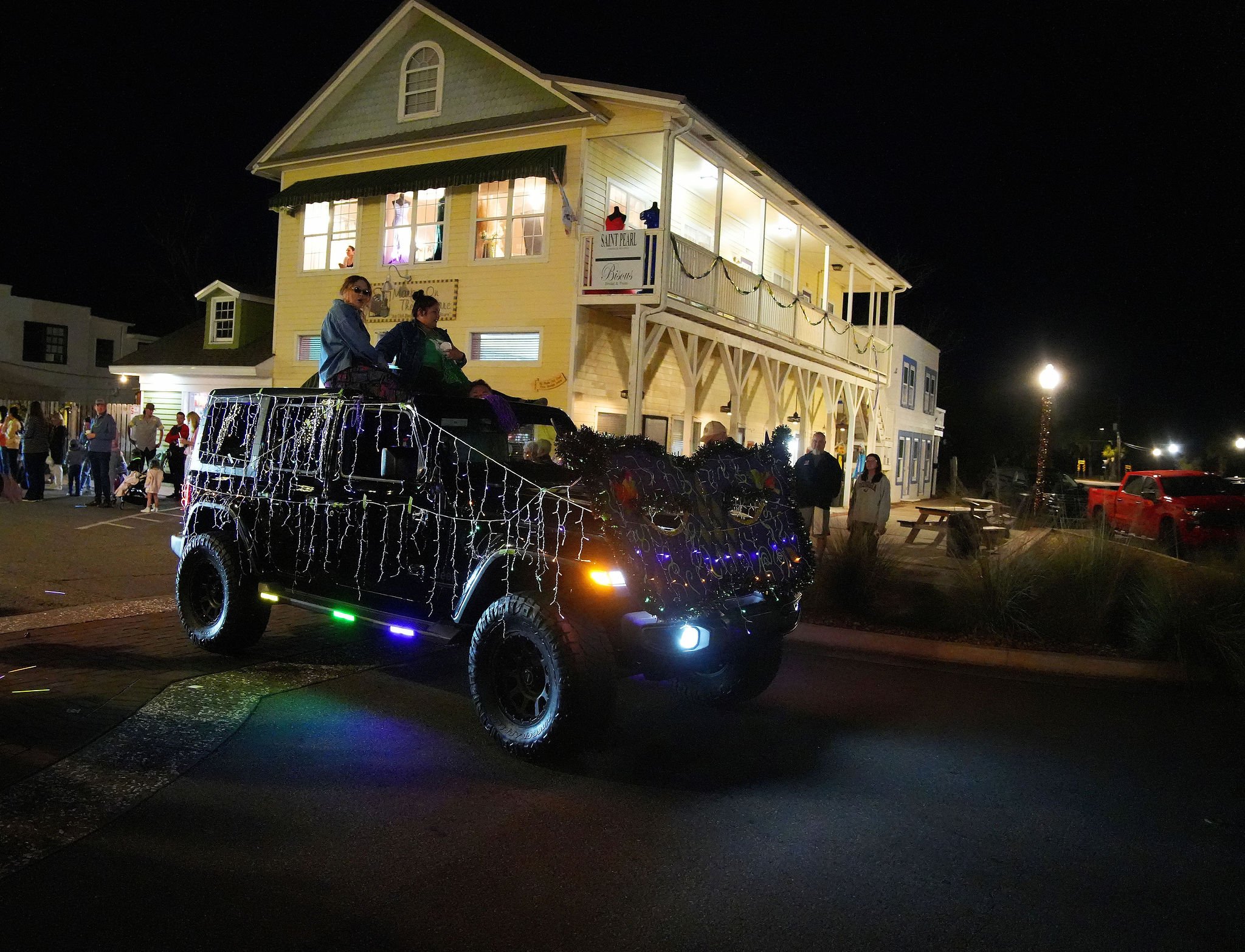 A black vehicle decorated with Christmas lights and ornaments, carrying two women with sunglasses on top, driving through a street at night during a parade, with a two-story yellow building illuminated in the background and groups of people watching.