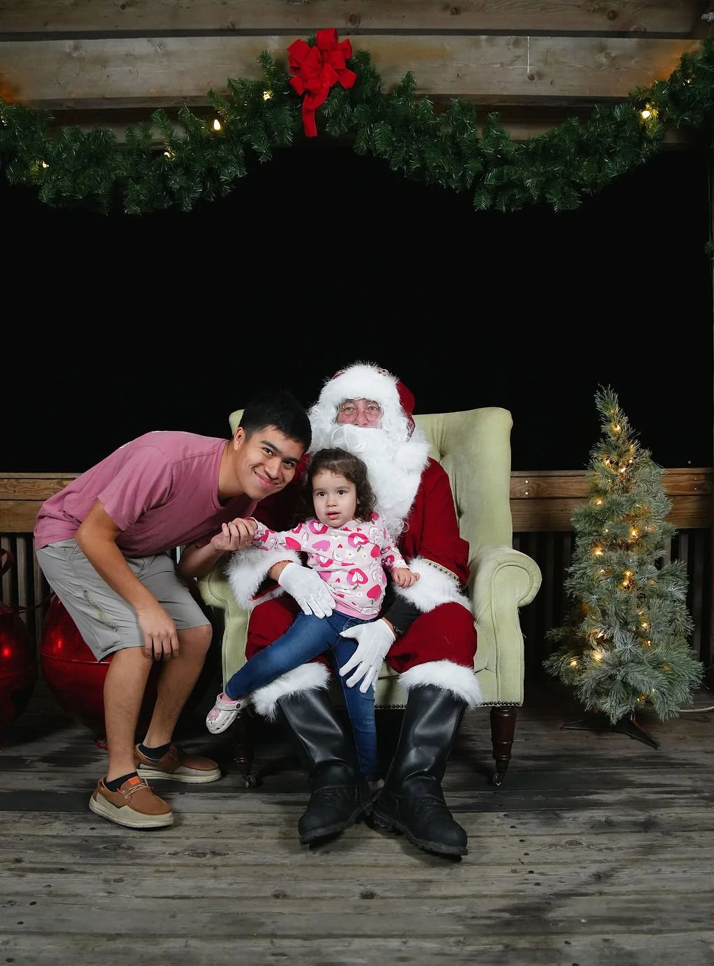 A young man, a little girl, and Santa Claus sitting in front of a black backdrop with Christmas decorations, including garland, a small Christmas tree, and red ornaments. The young man is leaning toward the girl, holding her hand, while Santa Claus s