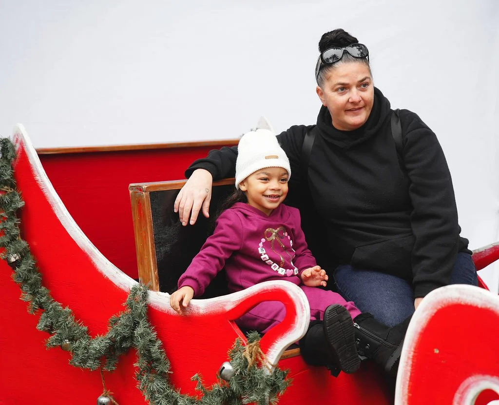 A woman and a young girl sitting in a Christmas-themed sleigh decorated with greenery and ornaments, smiling during a holiday event.