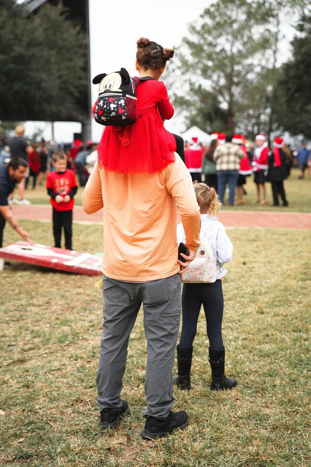 Person carrying a young girl on their shoulders at an outdoor holiday event with many people, some dressed in Santa costumes, in the background.
