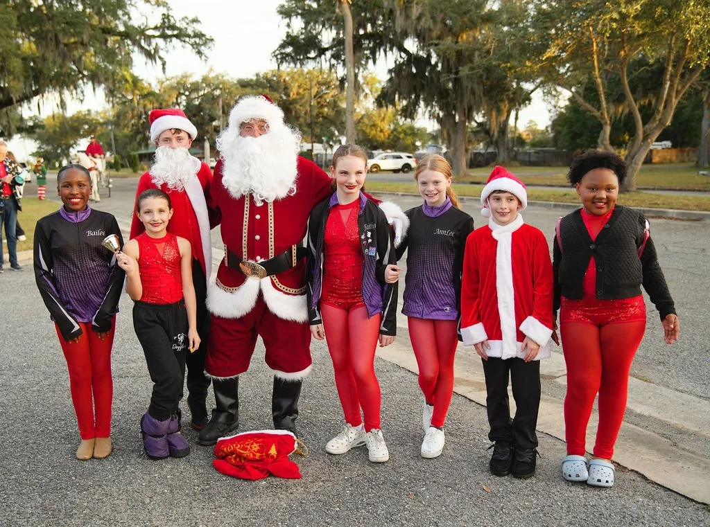 Group of children and adults dressed in Christmas costumes, including Santa Claus, posing outdoors on a scenic street with trees and parked cars.