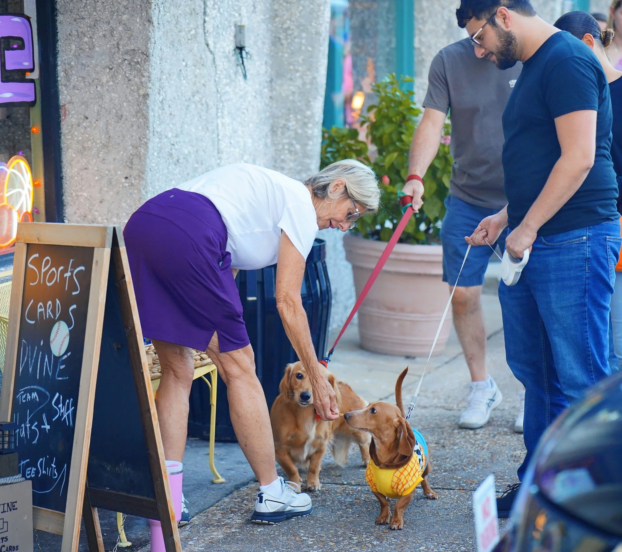 A woman and a man with dogs outside a store with a sign listing sports cards, playing cards, and T-shirts.