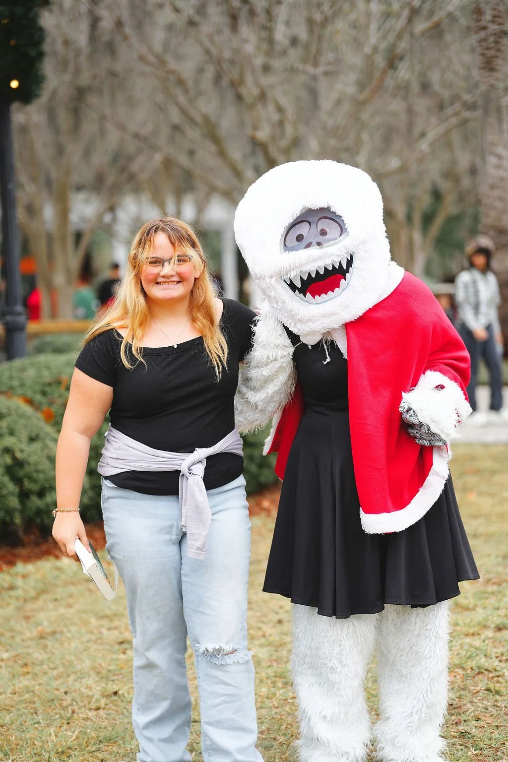 A woman smiling and standing next to a person in a snow monster costume dressed in a red cape and black dress, outdoors in a park with trees and other people in the background.