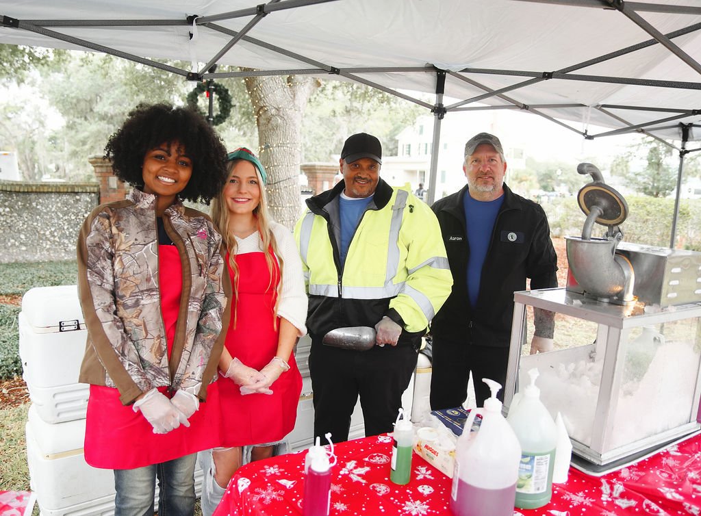 Four people standing behind a cotton candy machine at an outdoor event, smiling, with a red tablecloth decorated for Christmas, and bottles of supplies on the table.